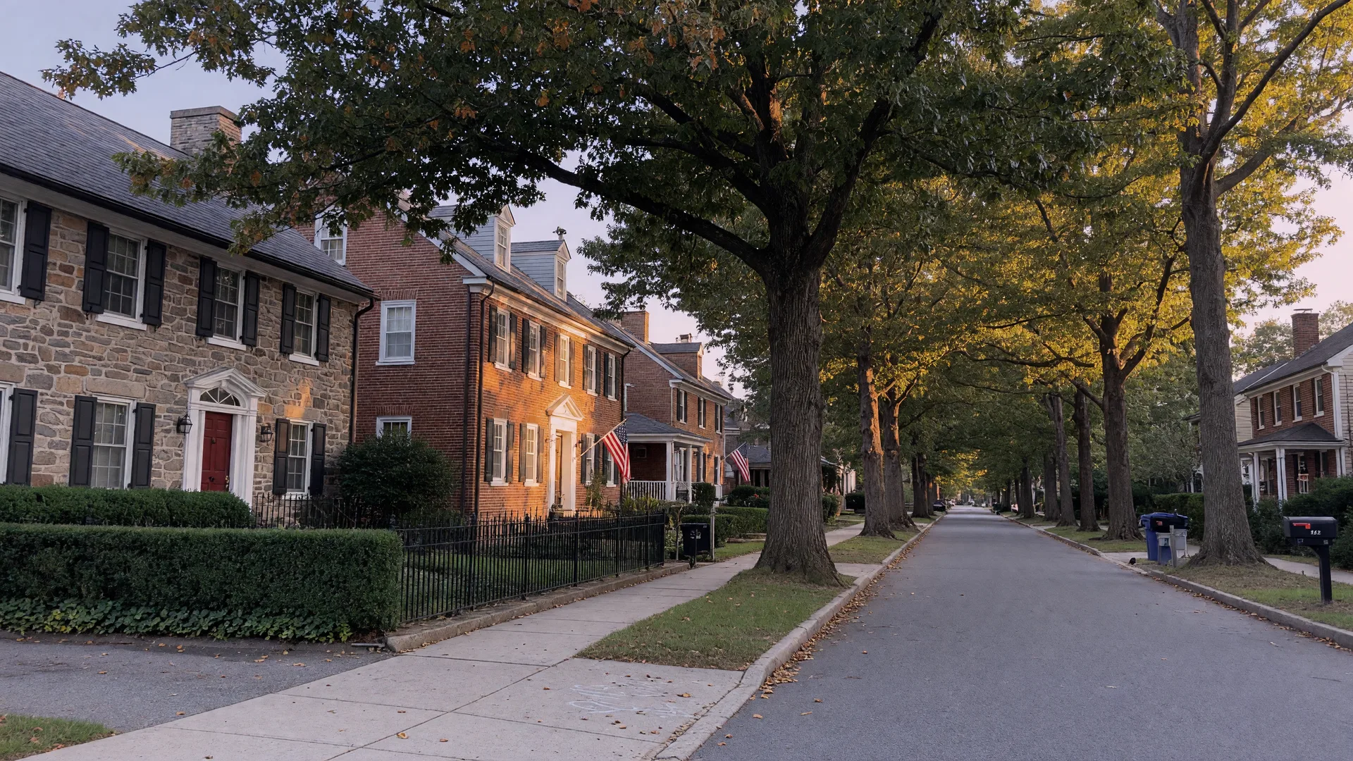 A tree-lined residential street in Rydal PA in mid-September with golden late-afternoon light across stone and brick homes under mature oaks with the first hints of autumn color