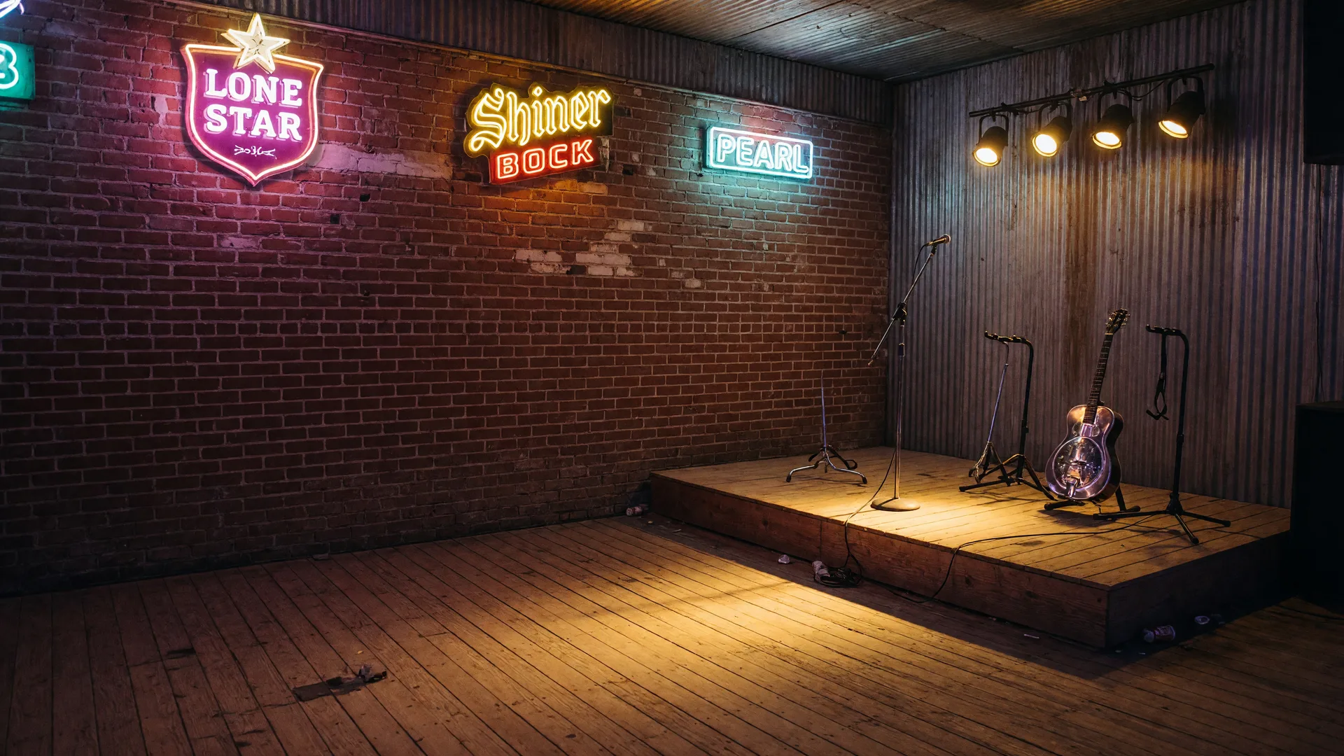 A classic Texas icehouse music venue at night with a weathered brick wall, neon beer signs, a small stage with guitars and a microphone, and warm tungsten light on worn wooden floors