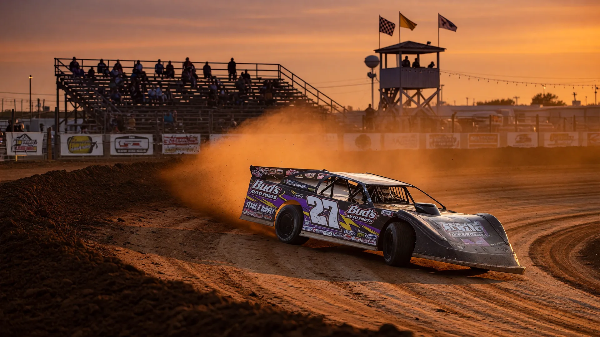 A dirt track race car kicks up dust on a high-banked clay oval during golden hour at a rural Northeast Texas racetrack