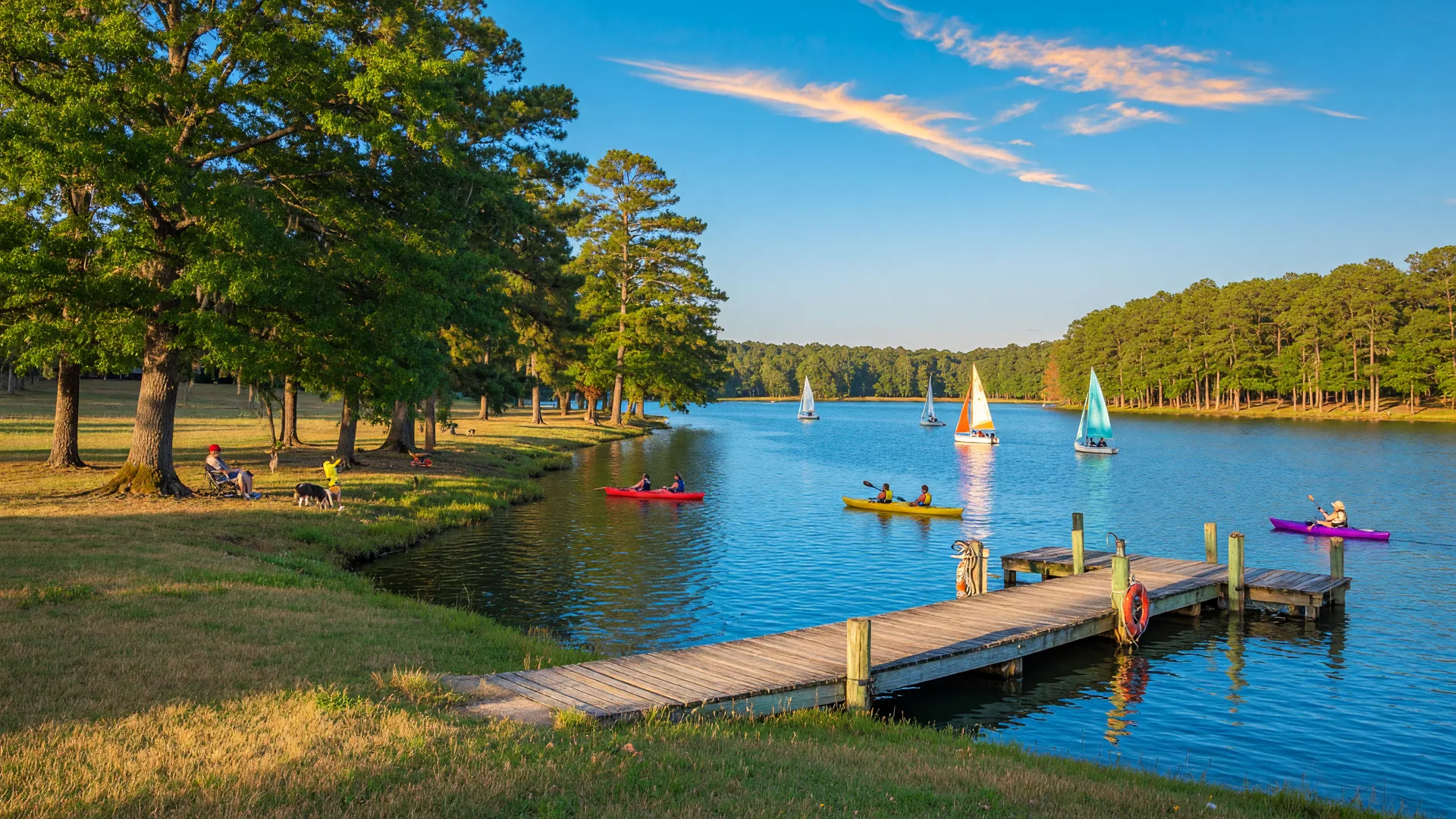 Lake Acworth on a sunny afternoon with calm blue water, sailboats, a wooden dock, and mature shoreline trees under a bright blue sky