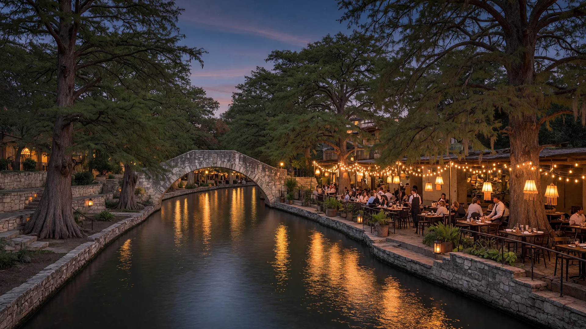 The San Antonio River Walk at dusk with cypress trees, an arched stone bridge, and glowing cafe lights on the water