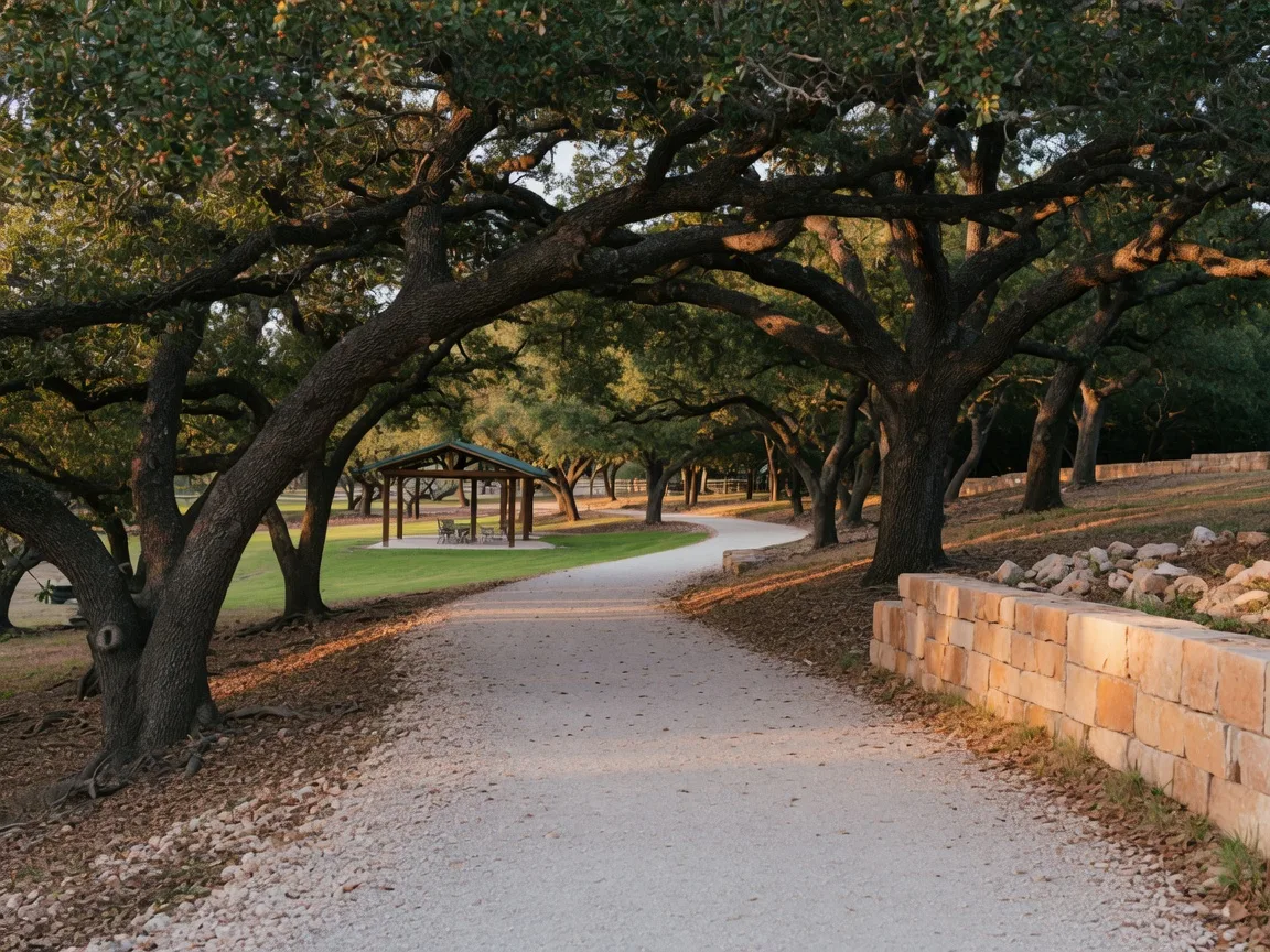 A crushed-granite walking trail winding under a canopy of mature live oaks with a small neighborhood park pavilion ahead in Fair Oaks Ranch, Texas