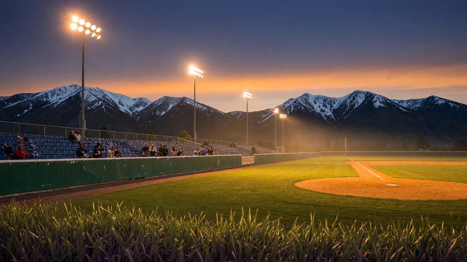 A minor league ballpark glowing under warm lights at dusk with snow-dusted mountain ridges beyond the outfield wall