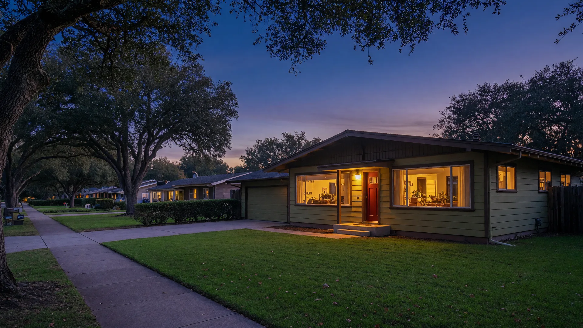 A mid-century ranch home in the East Bay hills at dusk with warm windows and a porch light glowing