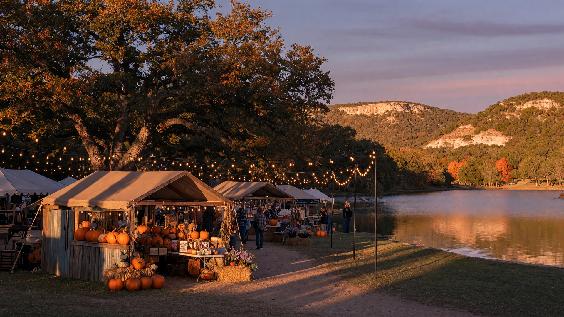 Texas Hill Country autumn festival evening, wooden market stalls with warm string lights strung between live oak trees, pumpkins and hay bales by a rustic stall, golden limestone hills and a calm lake under a dusk sky