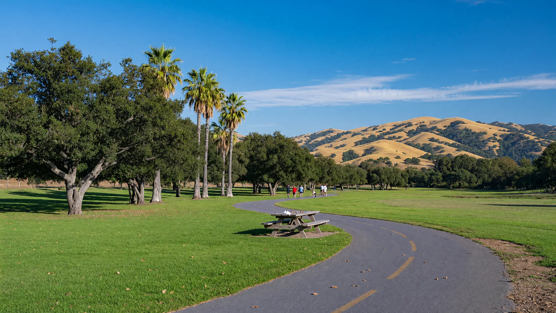 A sunny community park and walking trail in Murrieta with green lawn, oak and palm trees, and rolling foothills