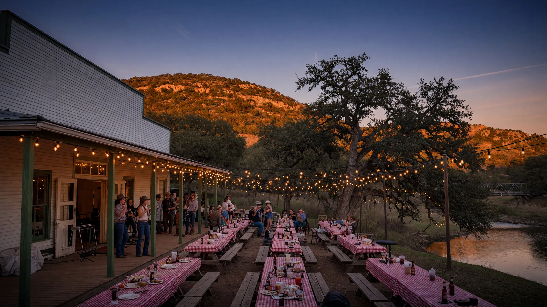 A festive autumn evening at Gruene Hall in the Texas Hill Country, warm string lights glowing on the dance hall porch, wooden picnic tables under live oaks, silhouetted guests enjoying live music, golden hills beyond