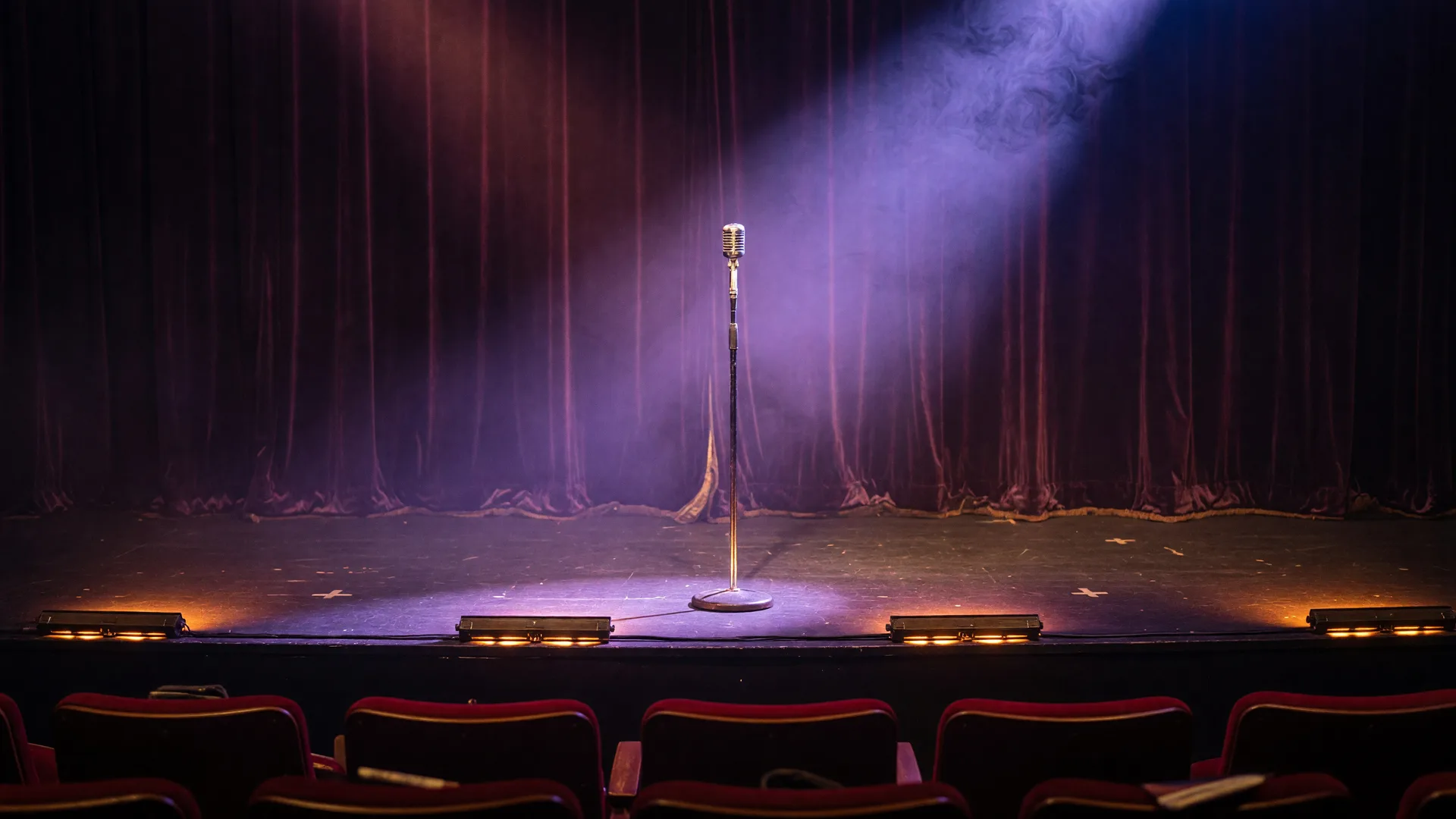 An empty comedy club stage with a single microphone under a warm purple spotlight cone and dark velvet curtains