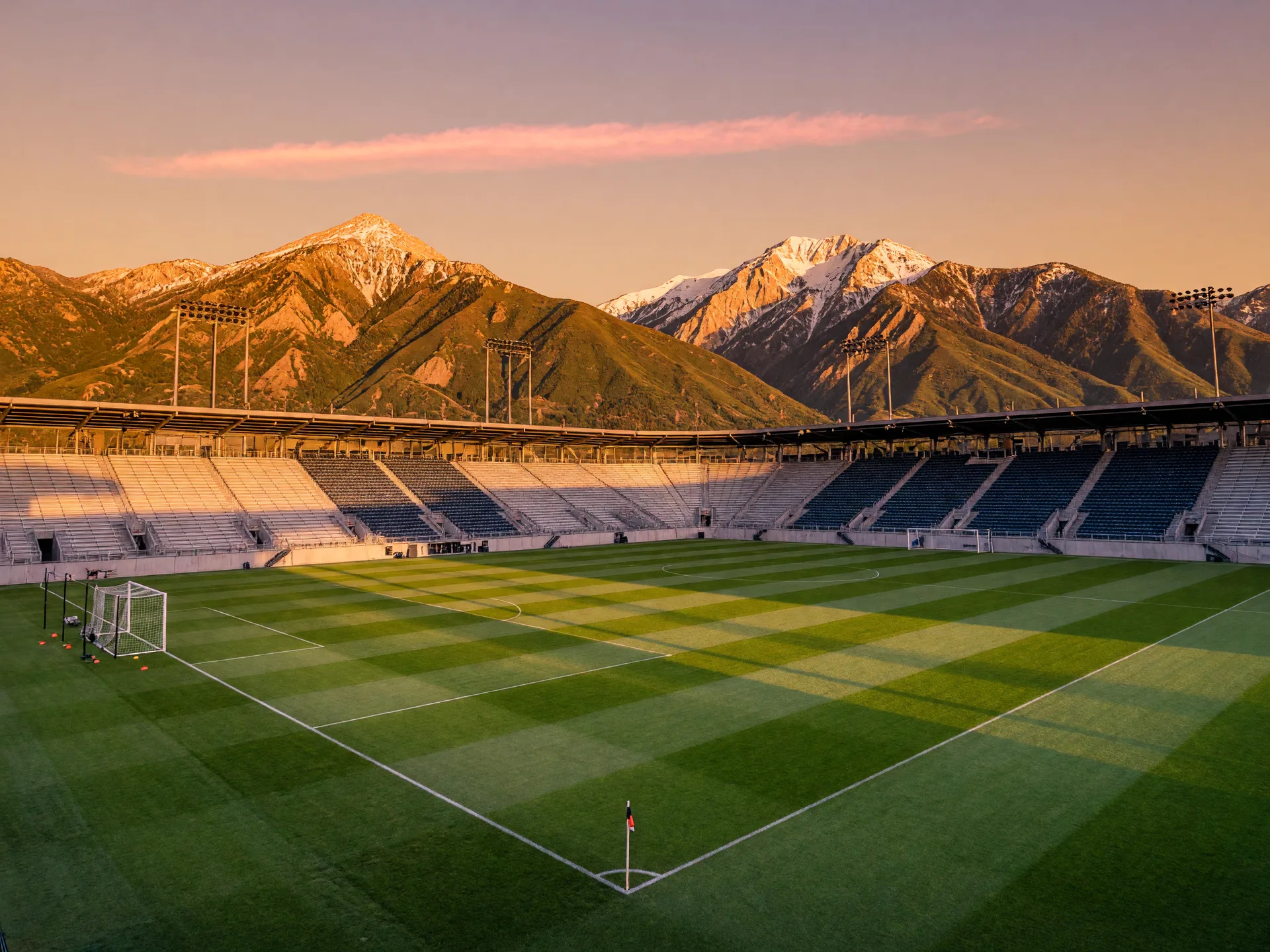 A soccer stadium bowl at golden hour with the snow-dusted Wasatch Mountains rising behind the stands