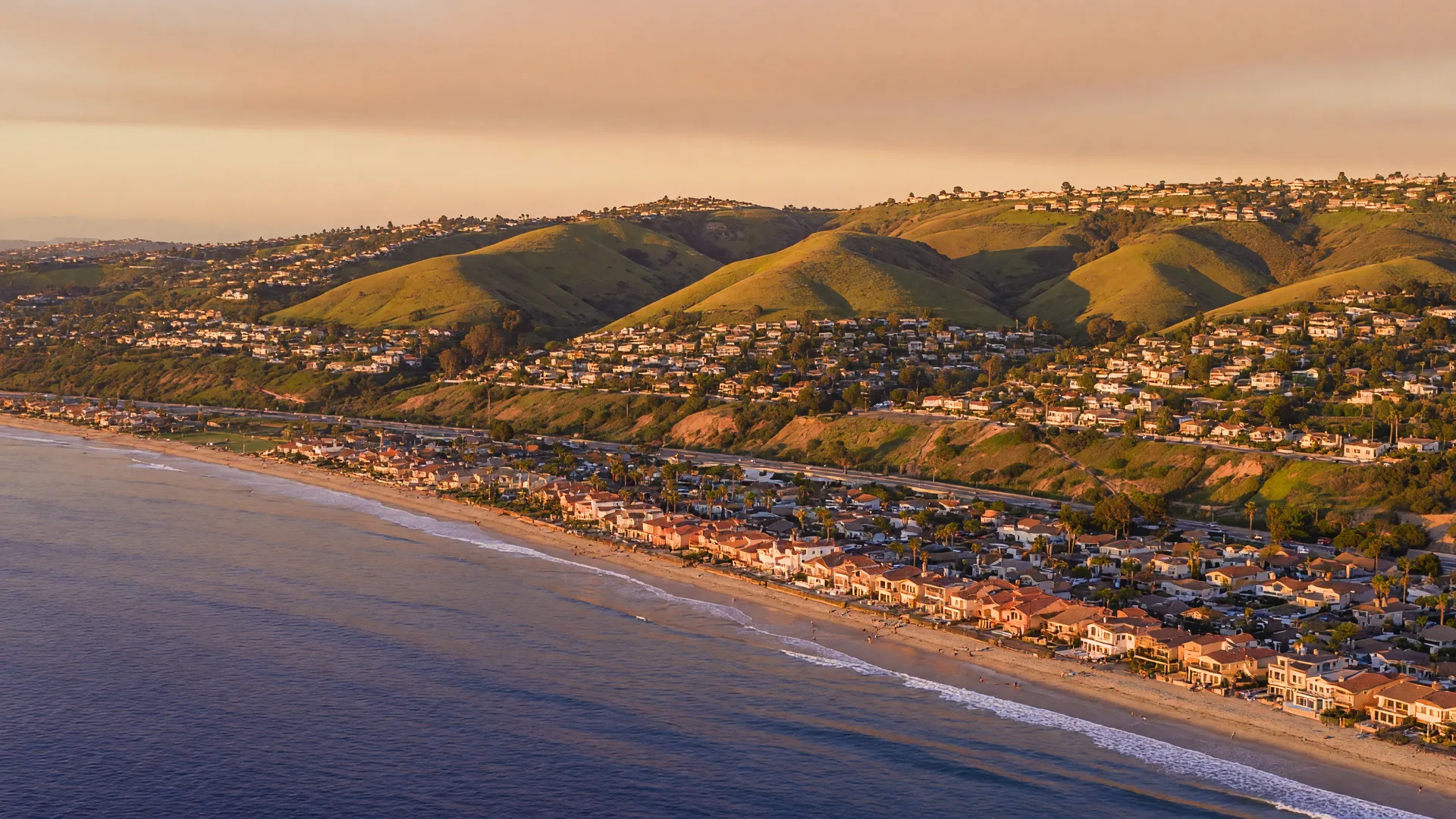An aerial view of North County San Diego at golden hour, coastal towns along the shore below green hills dotted with neighborhoods and the Pacific stretching to the horizon