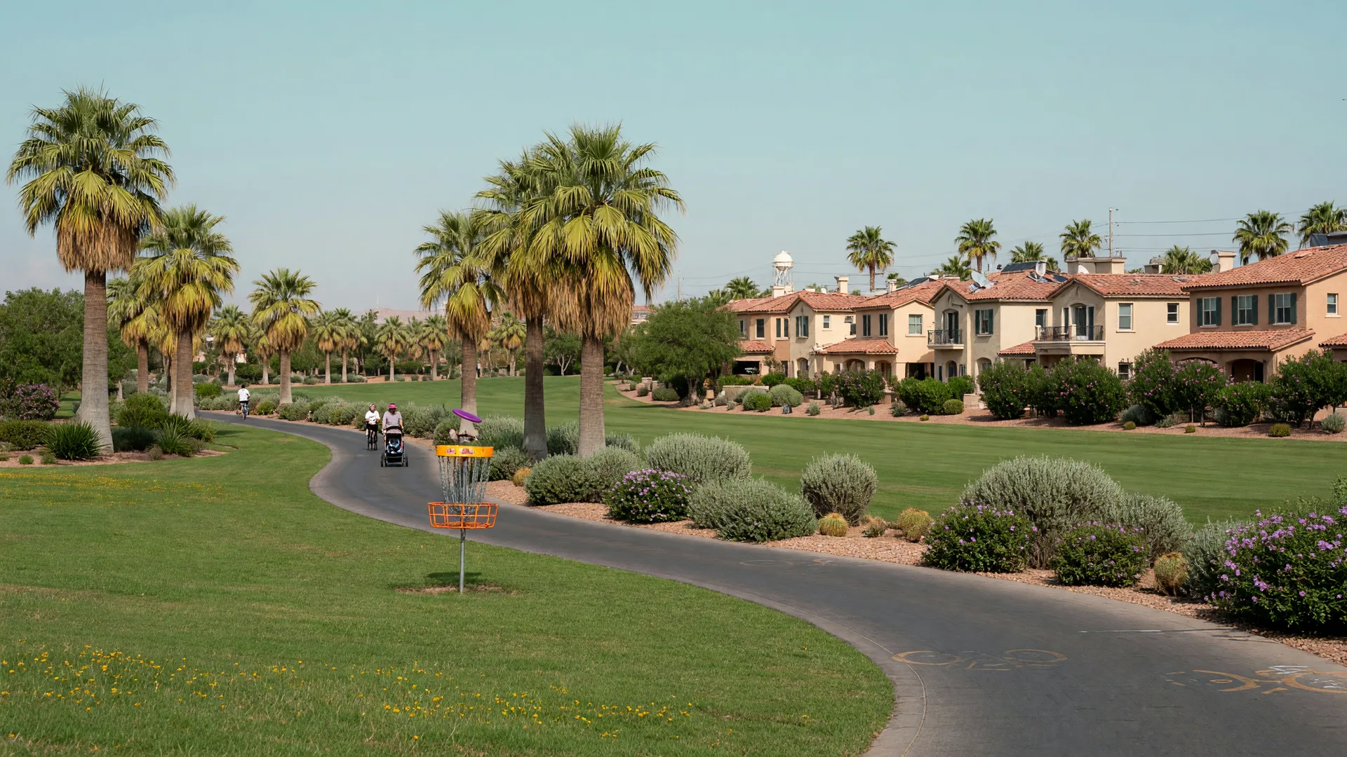 A greenbelt walking path through an established Las Vegas master-planned neighborhood with a disc golf basket and terra-cotta-roofed homes in the background