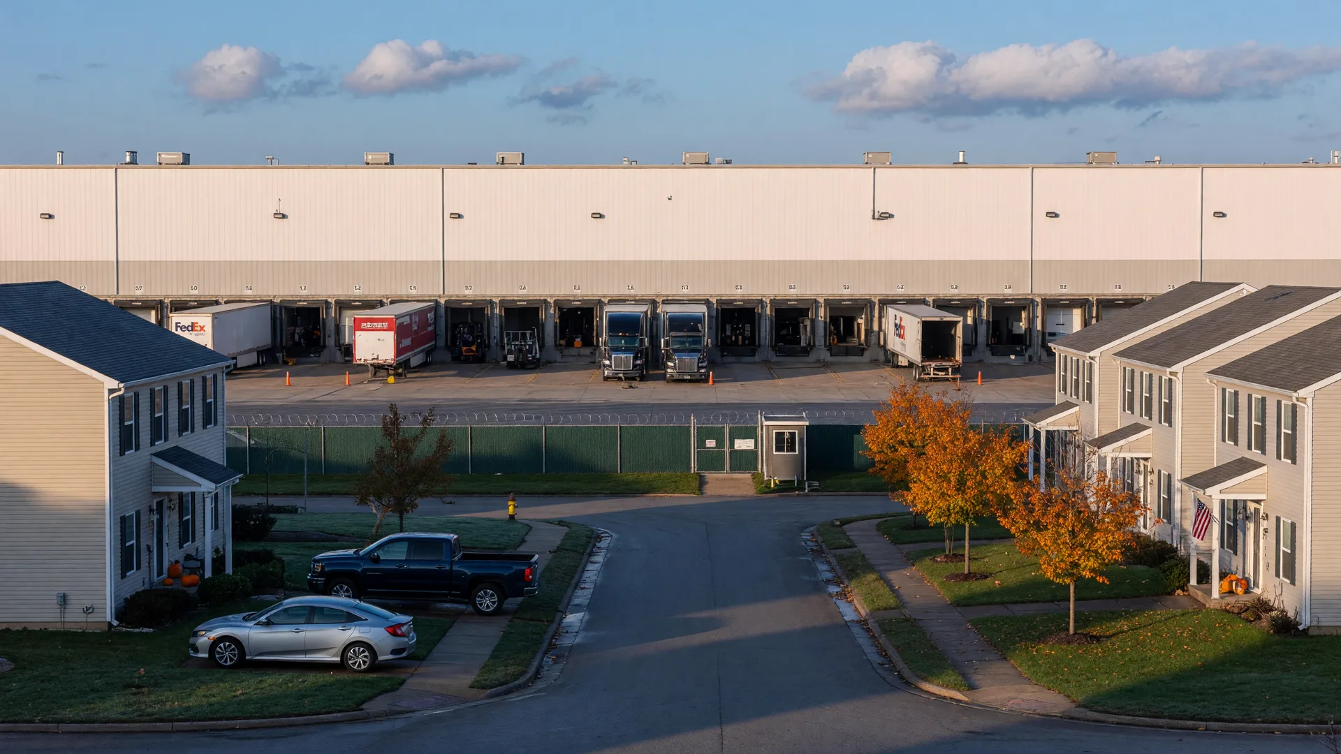 A long low white e-commerce fulfillment center with loading docks and parked semi trucks seen across a quiet suburban street in central Ohio, with newer two-story homes and curbside cars in the foreground and trees turning orange and gold