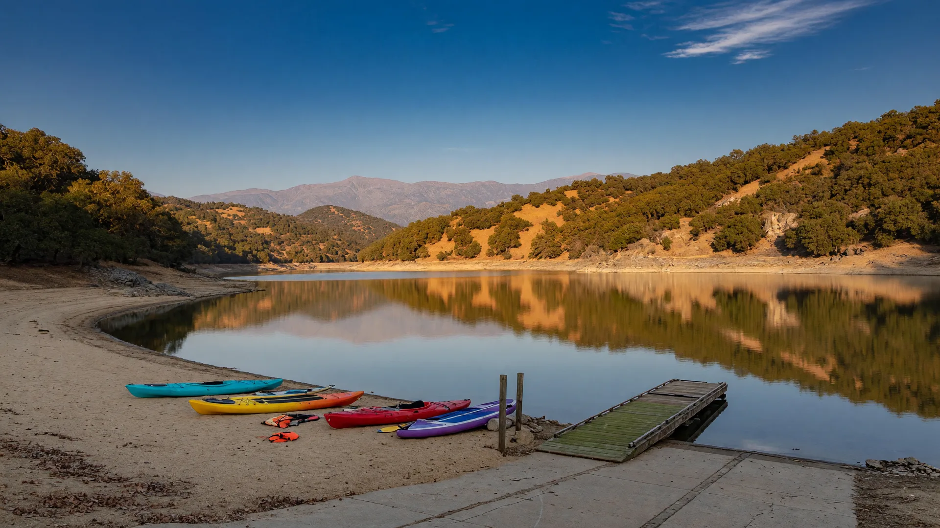 A calm Sierra-foothill reservoir in the Central Valley on a clear mid-October afternoon, golden oak-covered hillsides, a sandy swim beach with colorful kayaks and paddleboards pulled up on the sand, glassy water reflecting the amber hills, and a wooden boat launch ramp