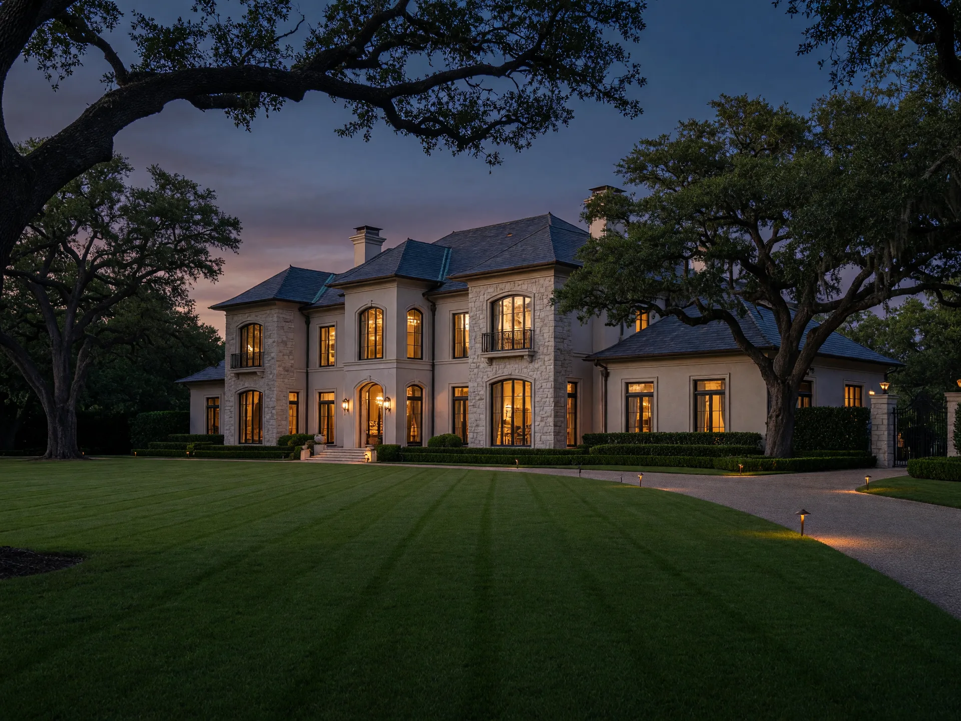 Dallas luxury estate at dusk with warm interior lights glowing through tall windows, live oaks framing the stone and stucco facade under a deep indigo sky