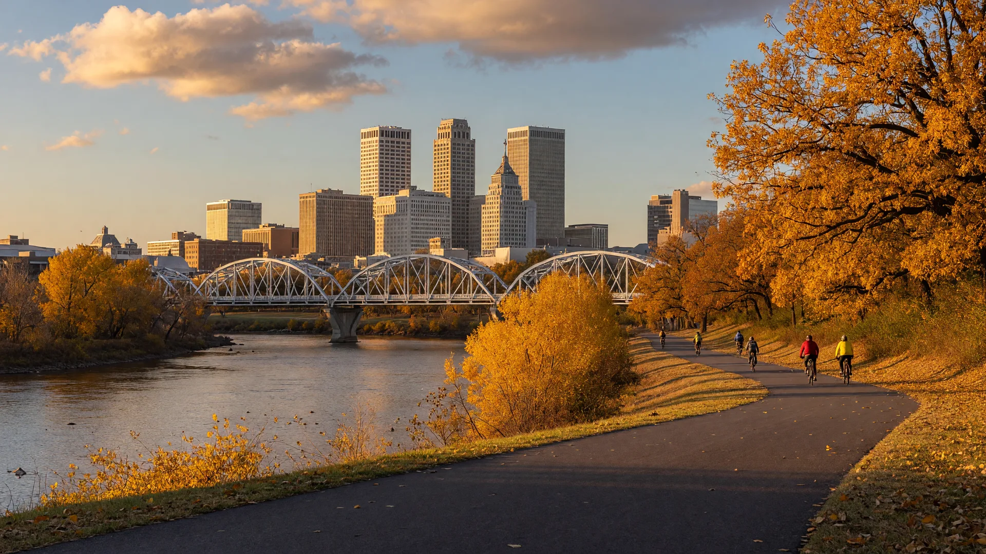A paved cycling trail curving along the Arkansas River in Tulsa on a golden October afternoon, with cyclists riding toward the downtown skyline and amber fall color along the riverbank