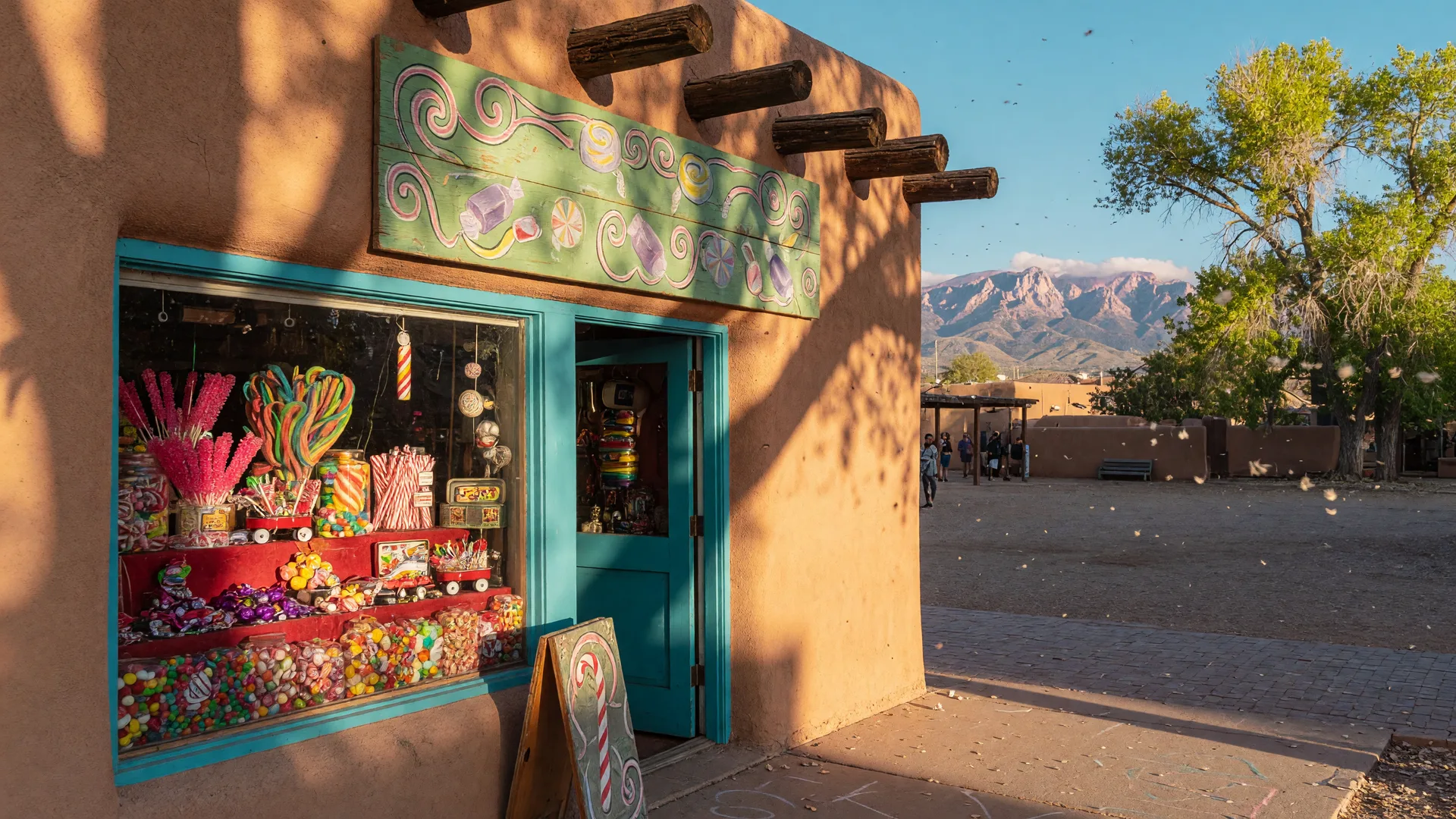 Candy shop storefront in Old Town Albuquerque glowing with warm morning light, adobe walls with wooden vigas and the Sandia Mountains in the distance