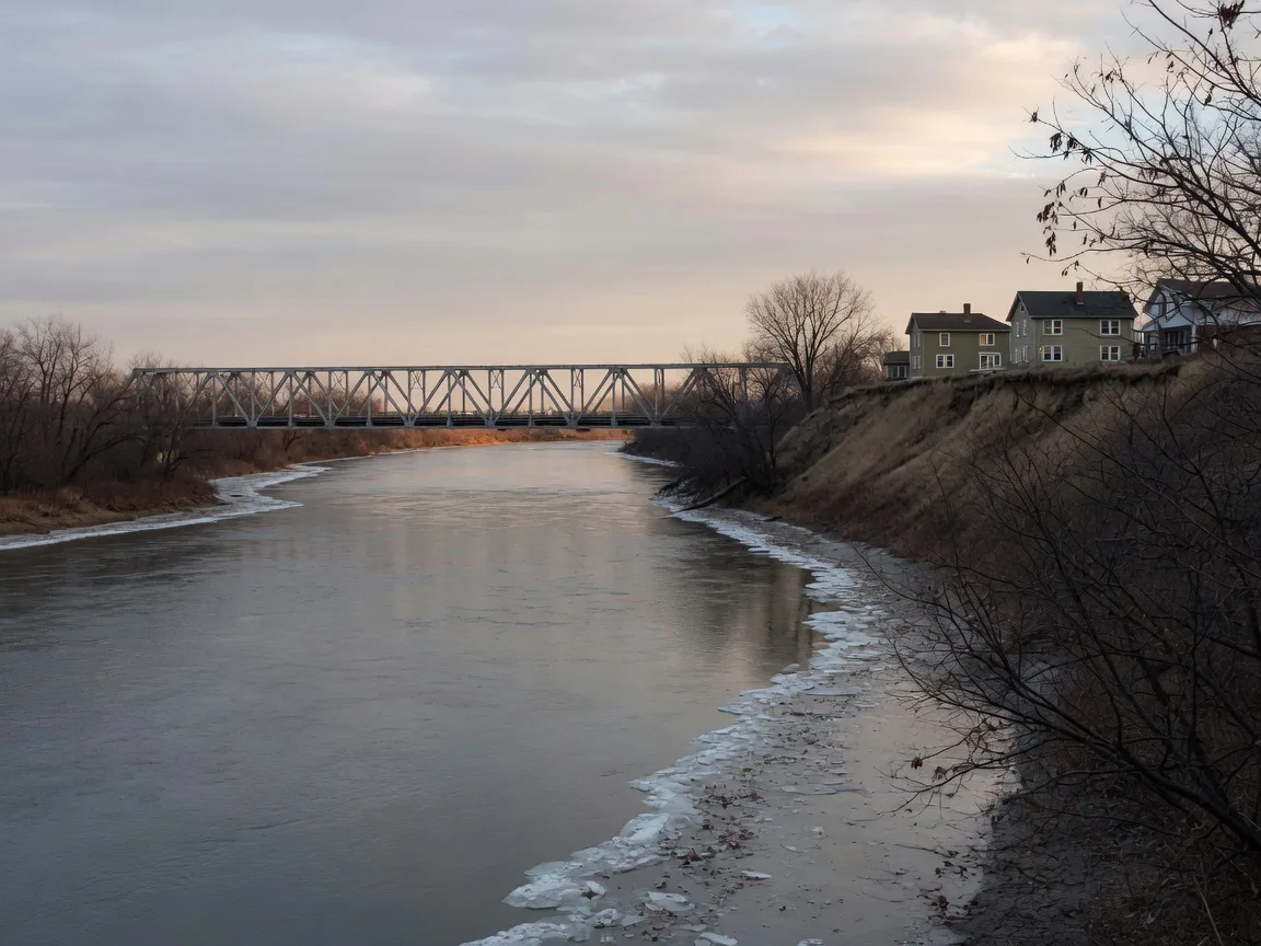 The Illinois River waterfront in Peoria in early December, with thin ice along the banks, bare trees, and a truss bridge in the distance under soft winter light