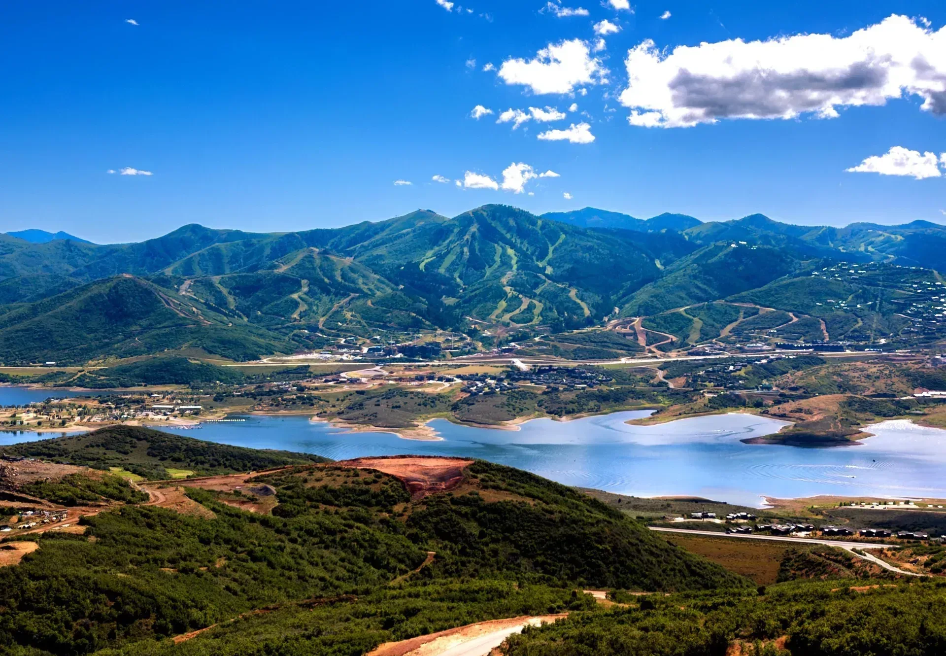Aerial view over a reservoir with ski slopes and mountain ridges beyond, under a bright blue sky with scattered clouds