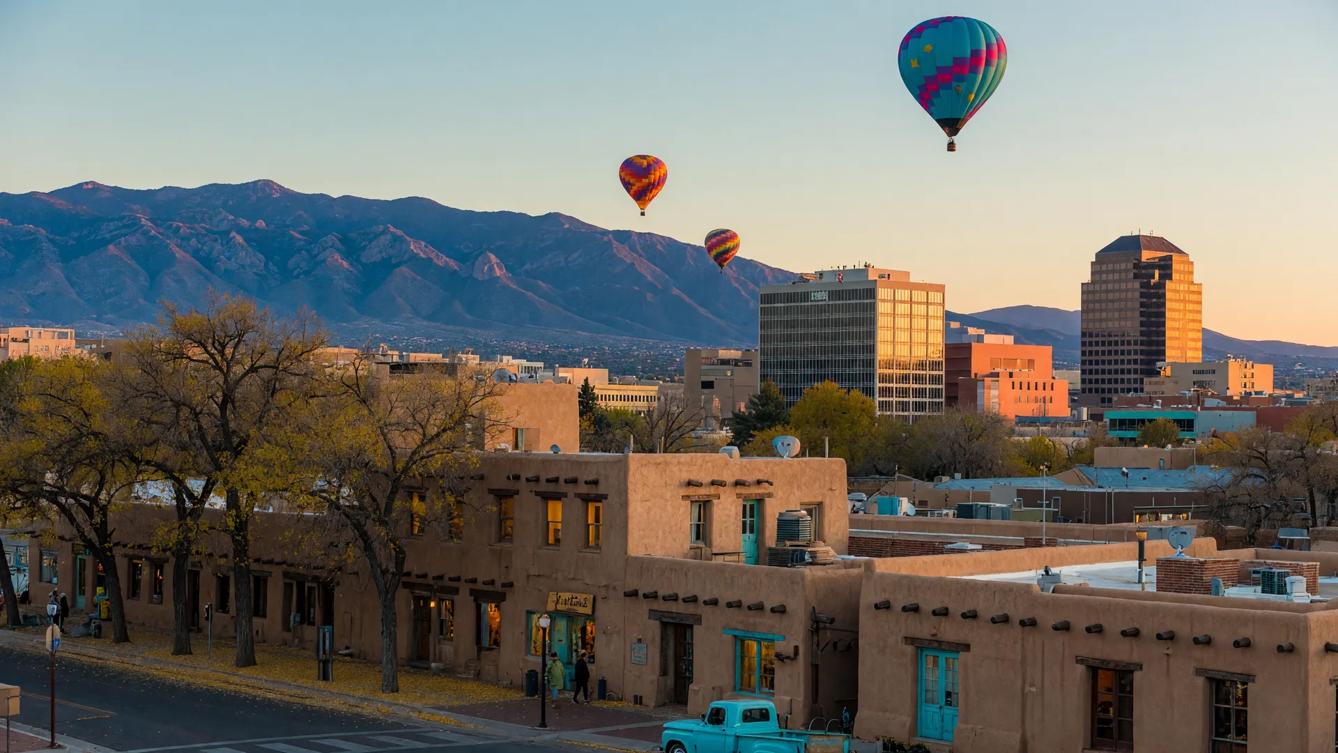 Downtown Albuquerque skyline at golden hour with adobe buildings in the foreground, hot air balloons in the sky, and the Sandia Mountains beyond