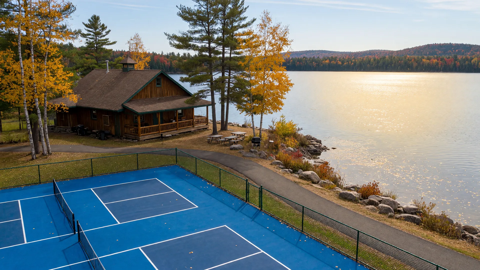 Fresh pickleball courts and a paved shoreline trail beside a wooden community center among pines and birch on a Minnesota lake