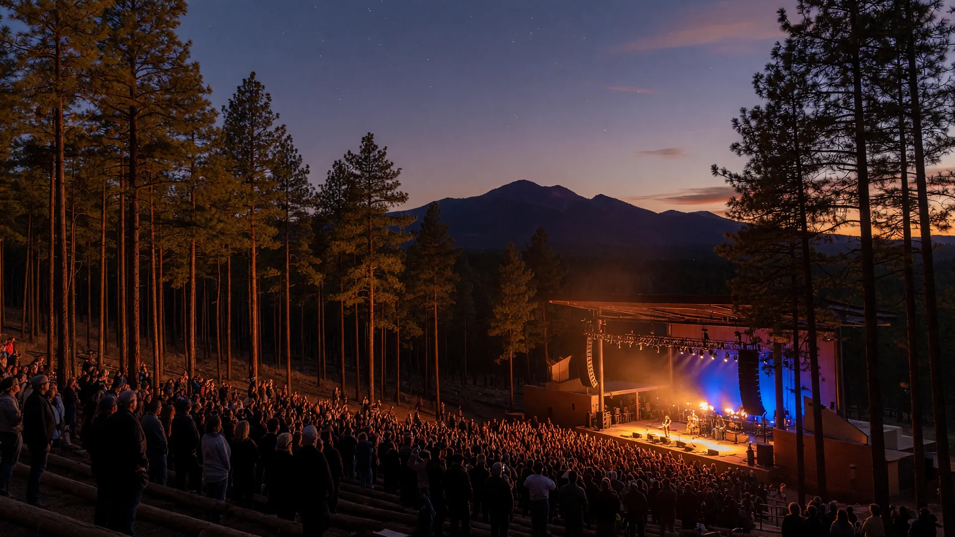 A concert crowd silhouetted against a warm amber-and-blue stage at an outdoor amphitheater in the ponderosa pines near Flagstaff at dusk