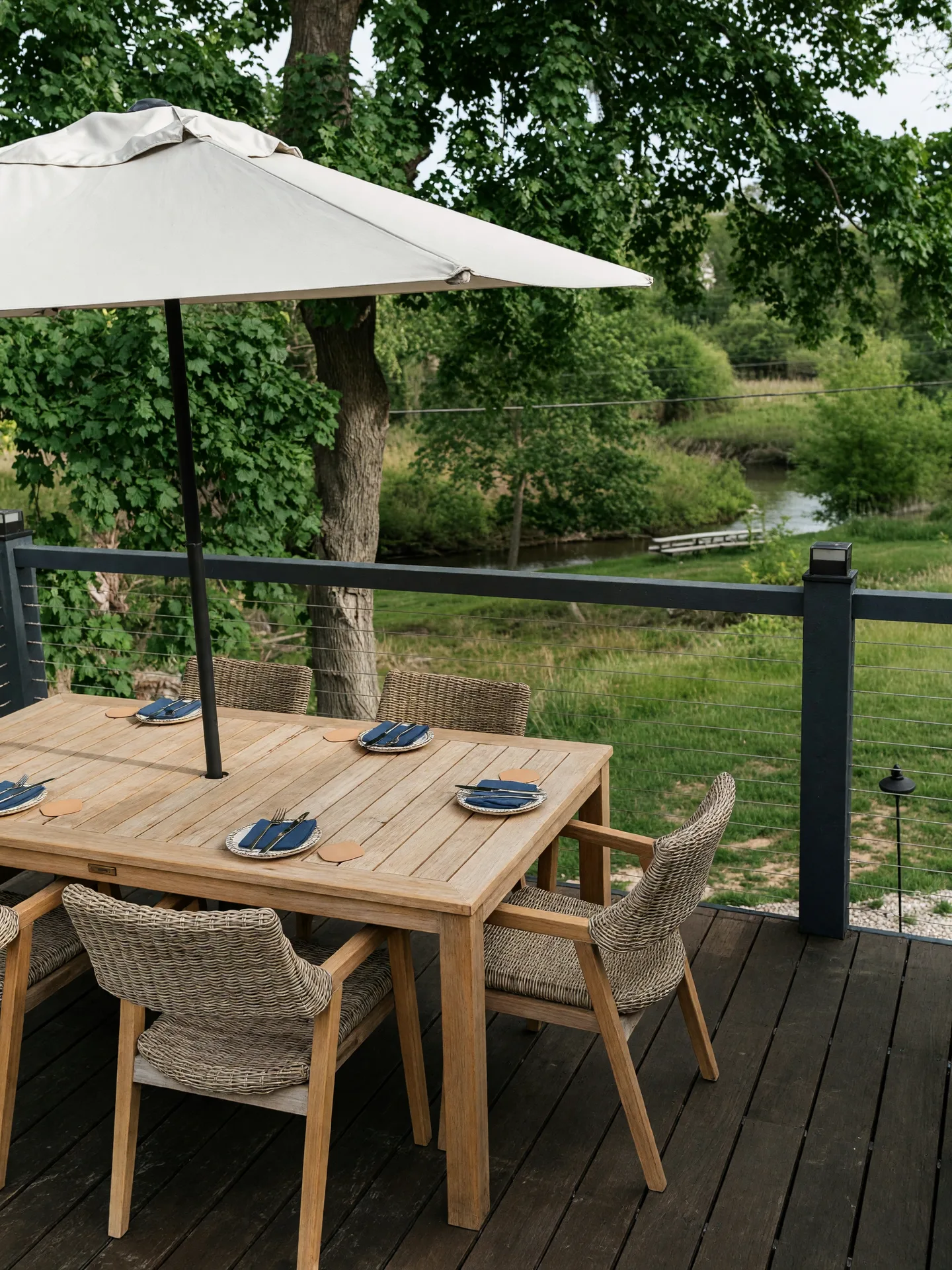 An outdoor wooden deck with a dining table, wicker chairs, and a large patio umbrella overlooking green landscaping