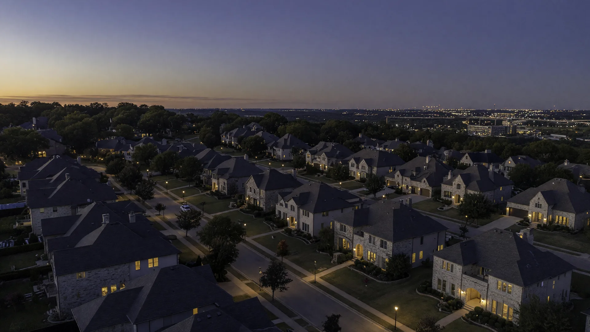 An aerial dusk view of a Tulsa Metro suburb, charcoal-roofed homes with warm golden window lights along tree-lined streets beneath a deep navy evening sky