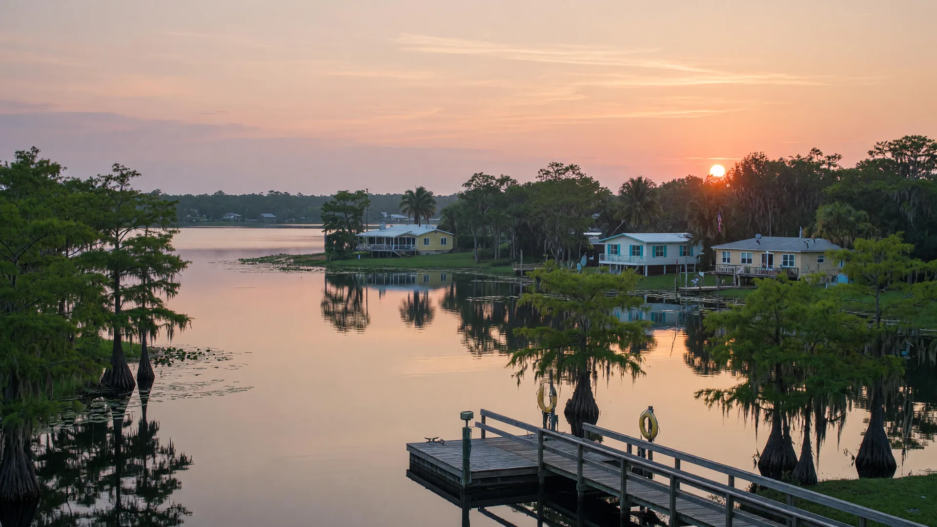 Kissimmee Florida chain of lakes at sunrise with cypress trees, a wooden dock over calm water, and homes visible along the treeline