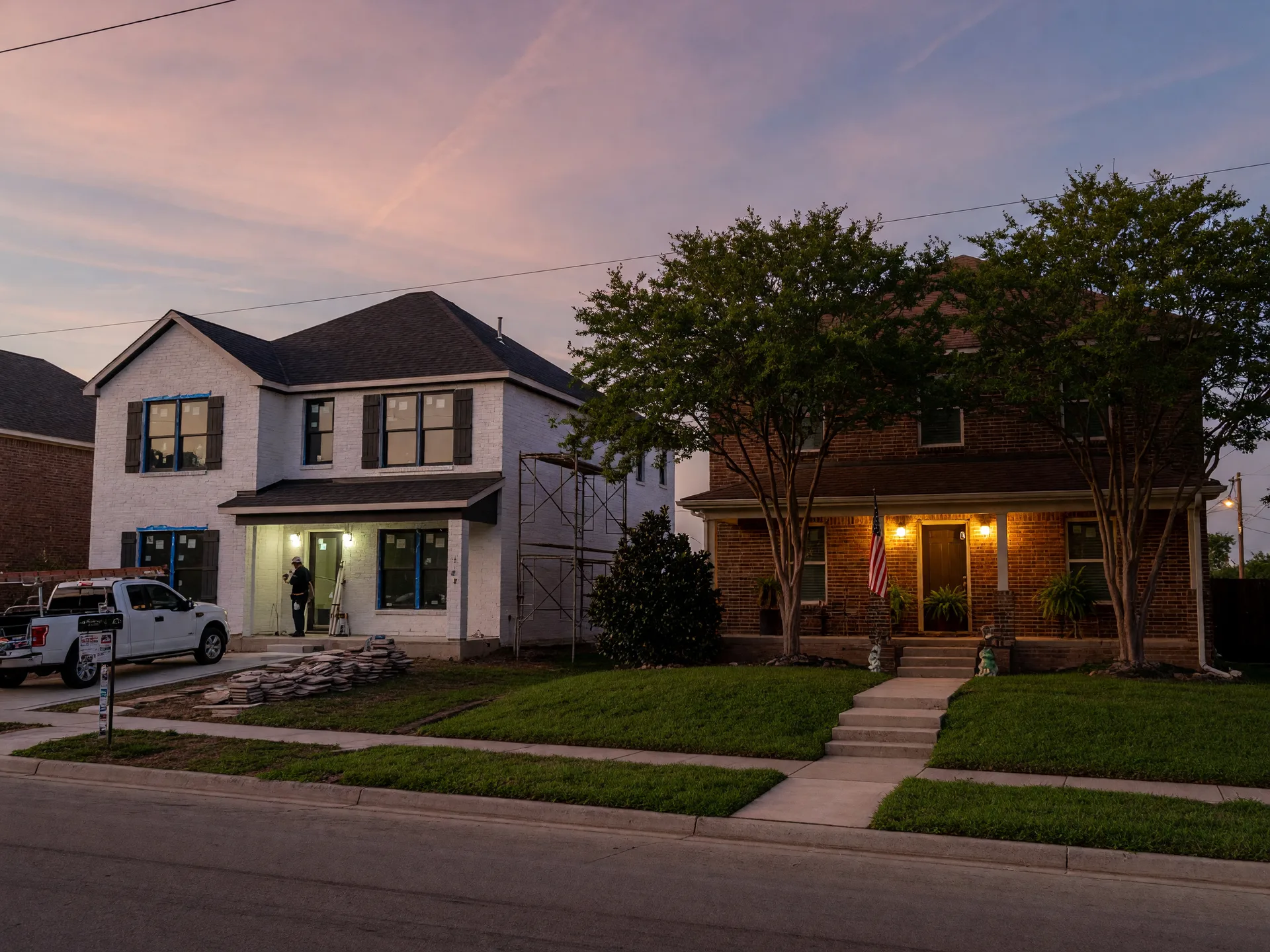 A new white-brick construction home beside an established brick resale home with mature trees on a quiet North Texas street at dusk