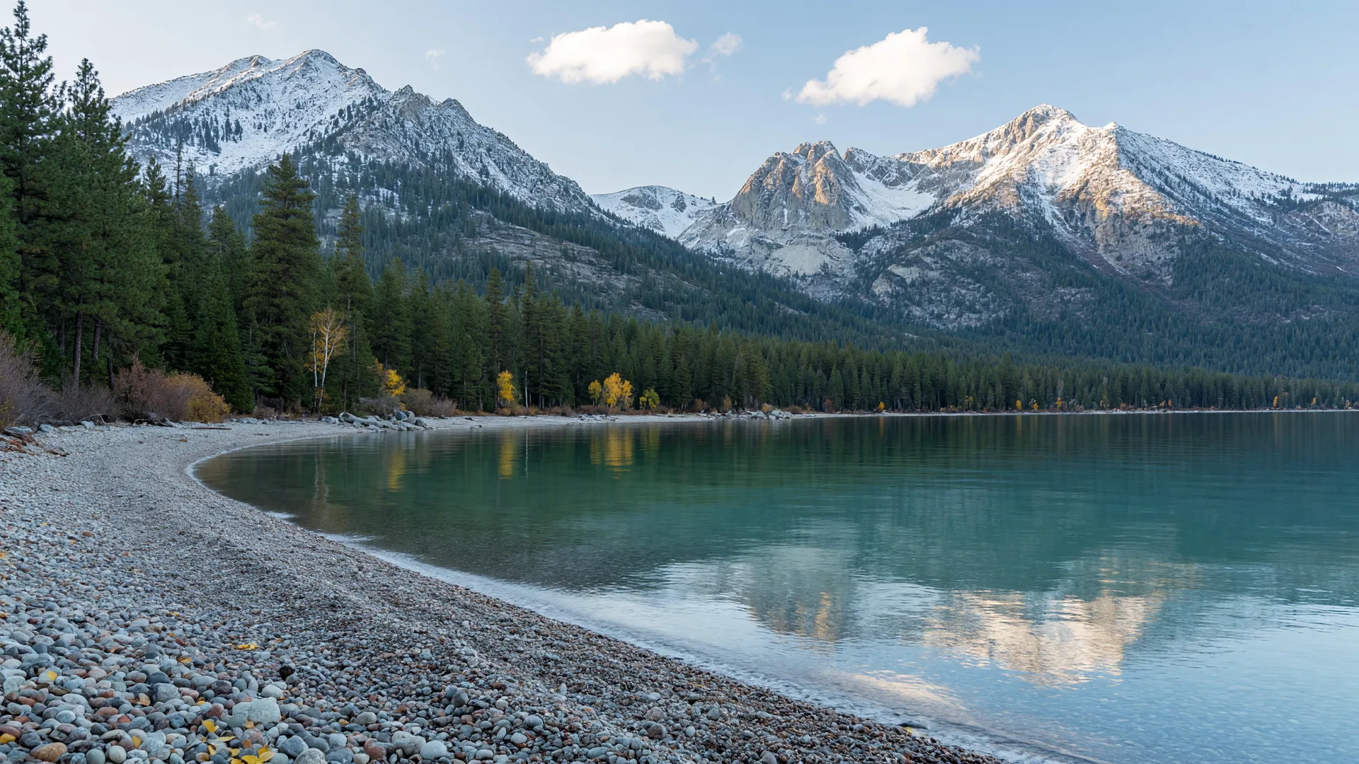 Lake Tahoe shoreline in early autumn with calm turquoise water, a pebbled beach, pine forest, and a dusting of snow on the granite peaks above.
