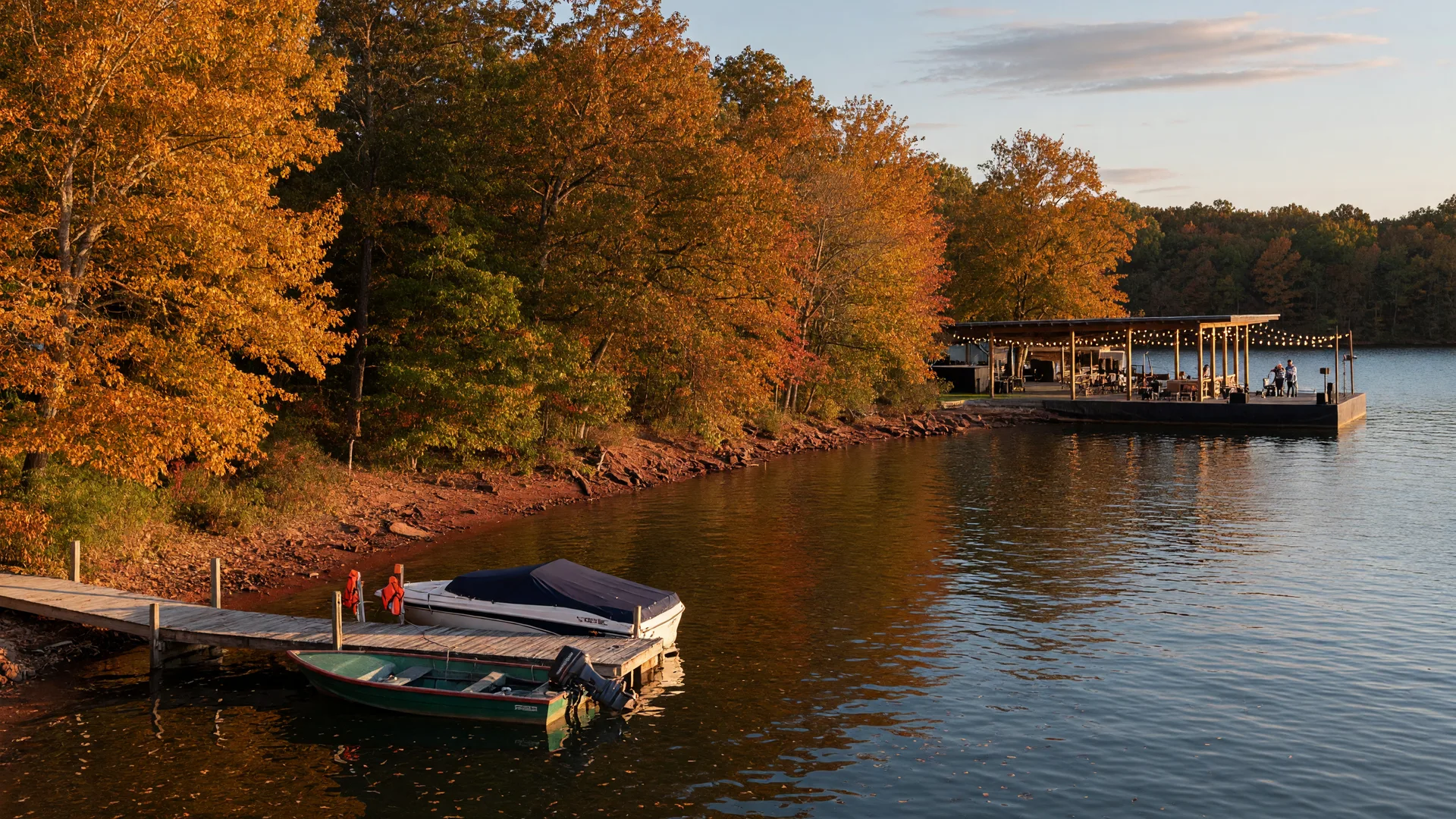A mid-October cove on Lake Norman with golden autumn foliage along the shoreline, a wooden dock with moored boats, and a string-lit lakeside venue with a small stage in the distance in warm afternoon light
