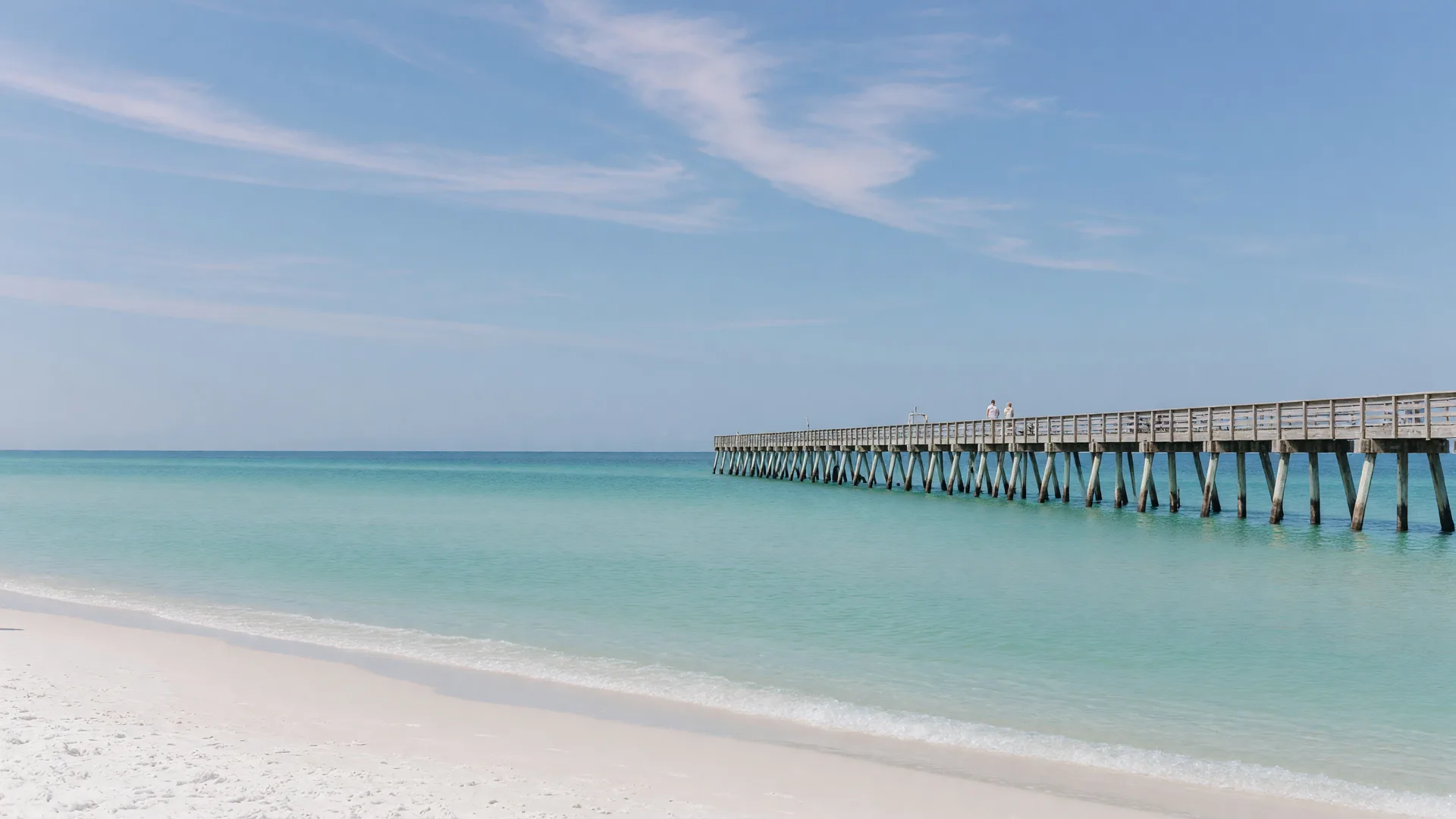 Pensacola Beach's white sand and turquoise Gulf water with a wooden pier reaching into the distance on a clear morning.