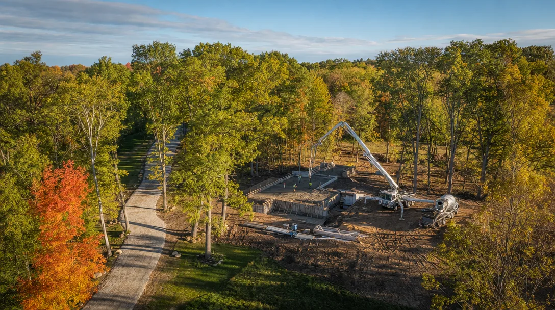Aerial view of a custom home construction site on a wooded one-acre lot in Southeast Michigan, showing a foundation pour among preserved mature trees