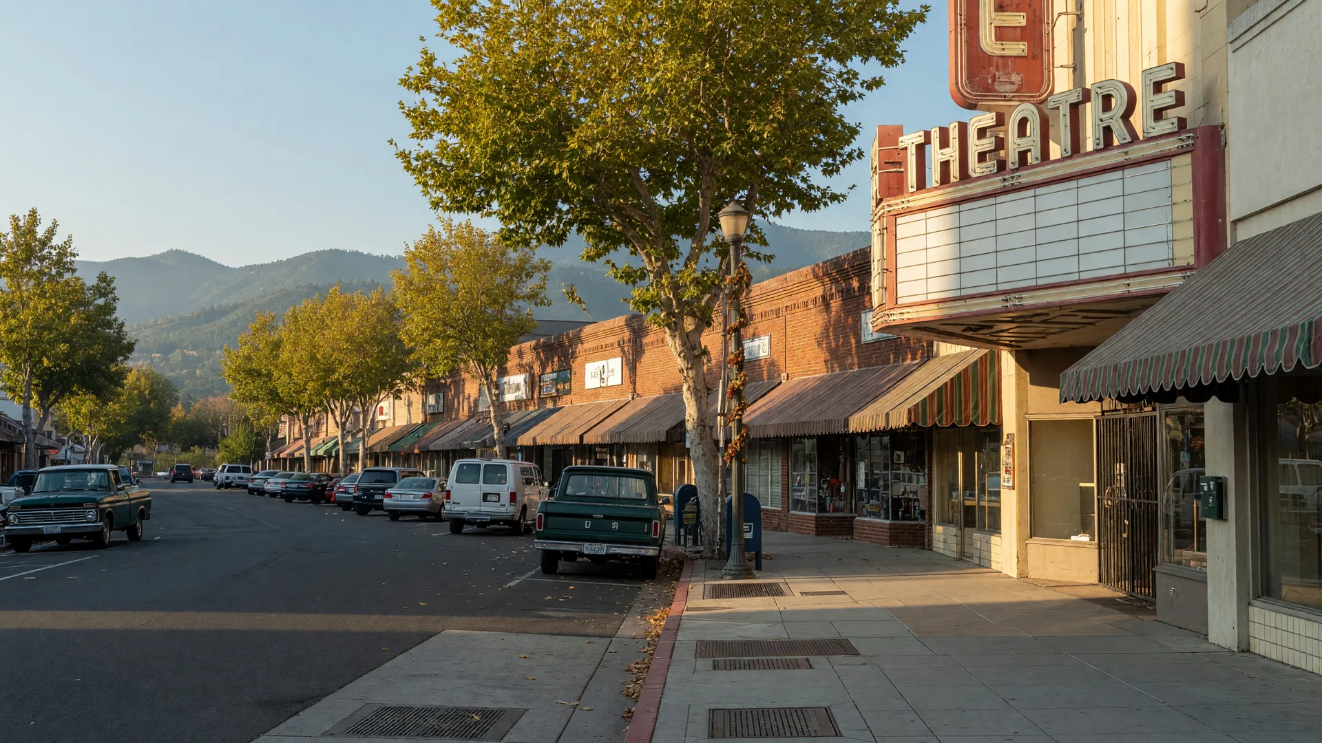 A small Central Valley downtown street in Merced County on a clear October morning, brick storefronts with awnings, a restored theater marquee, plane trees and parked cars, and the Sierra Nevada foothills on the horizon