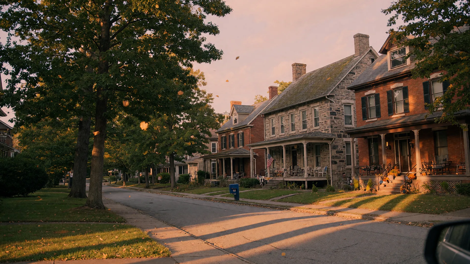 A tree-lined residential street in Rydal PA in mid-September with golden late-afternoon light across stone and brick homes under mature oaks with the first hints of autumn color