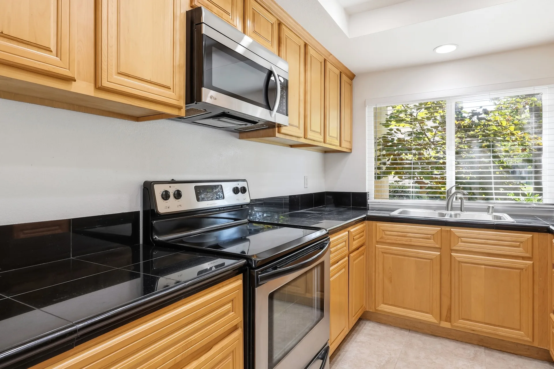 Galley kitchen at 8 Silverwood with light oak cabinetry, black tile counters, stainless steel appliances, and a window over foliage