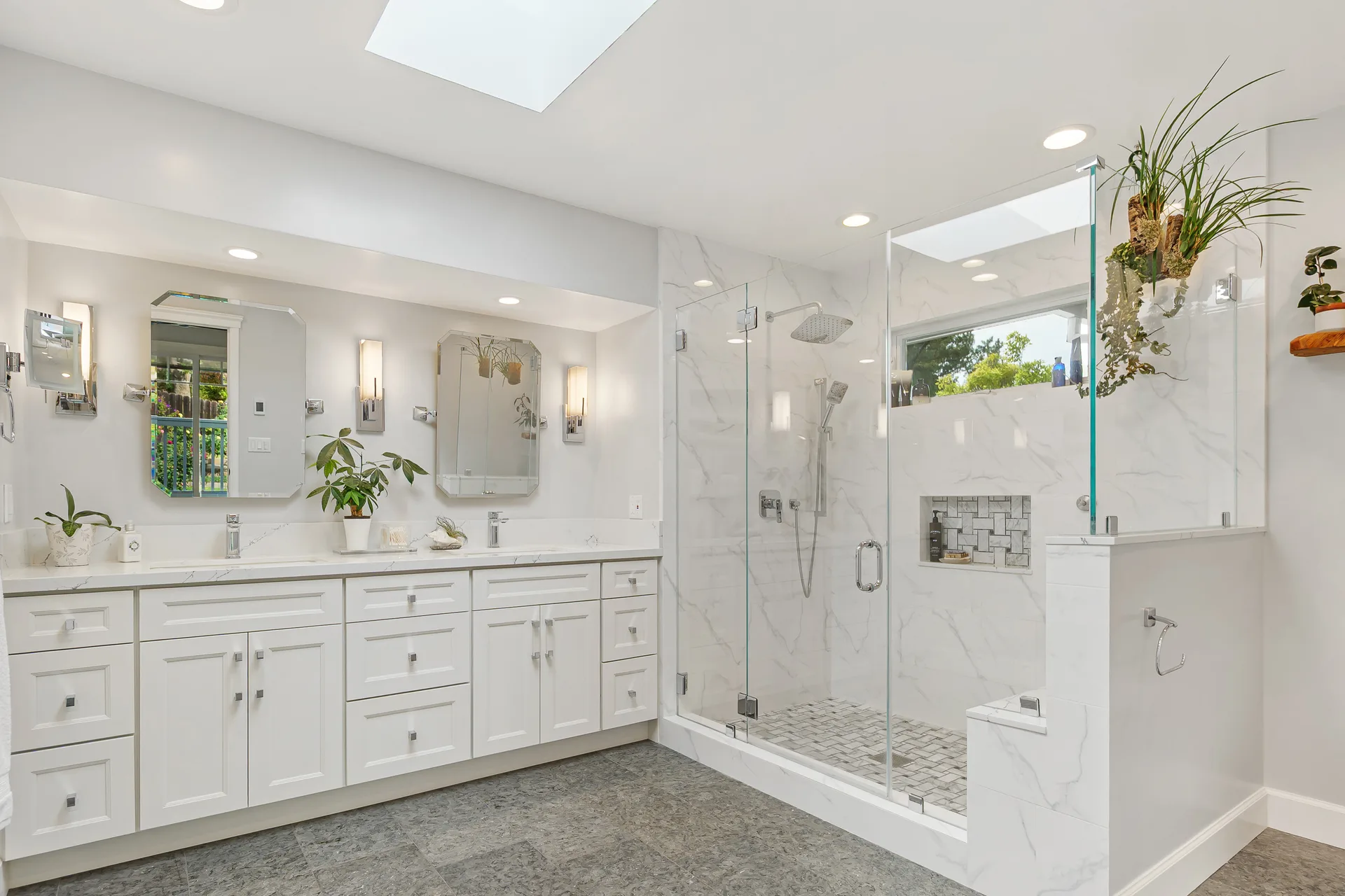 Renovated spa-inspired primary bathroom with double vanity, marble-look tile, glass shower, and skylight