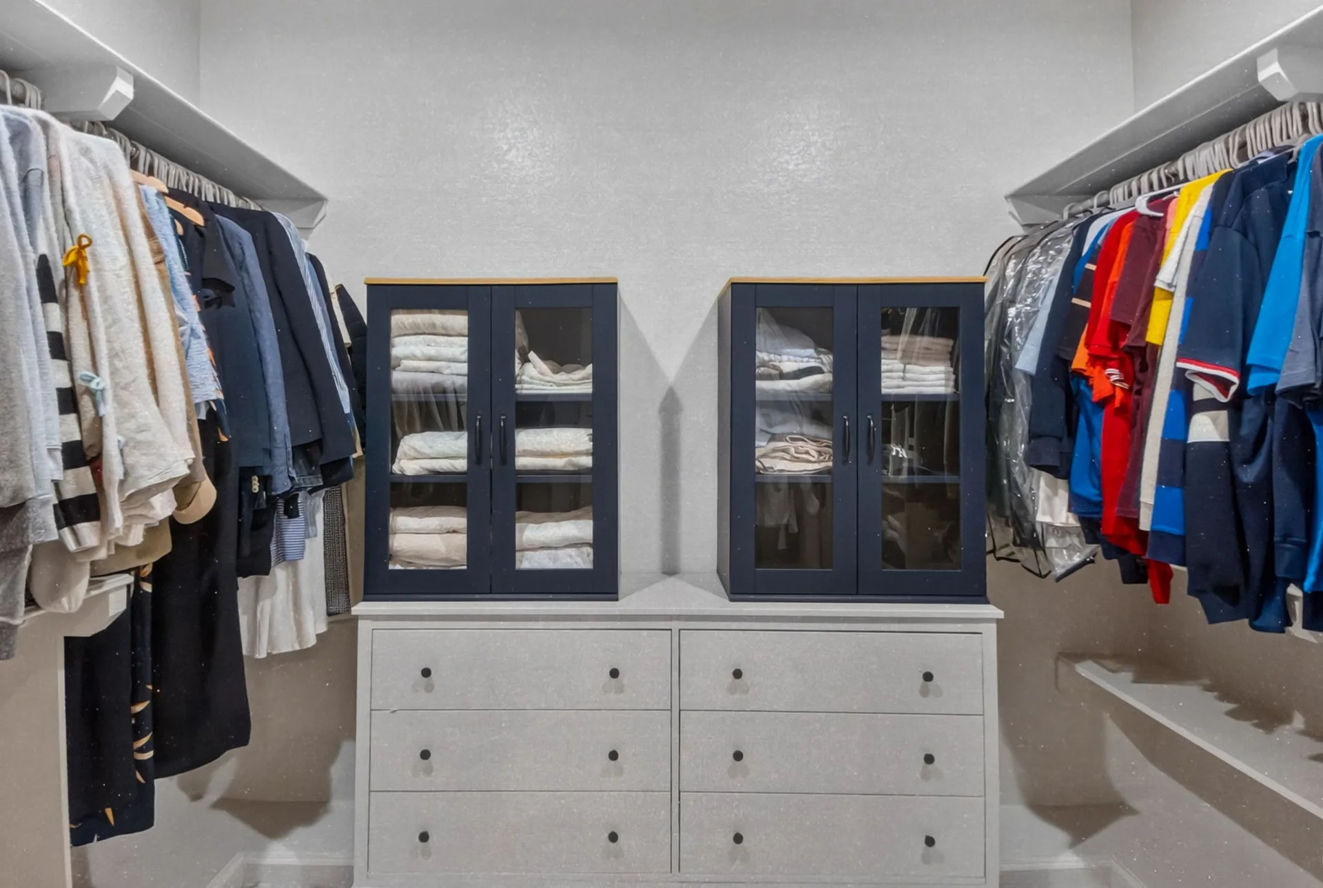 Custom walk-in primary closet with navy glass-front upper cabinets and a white dresser base