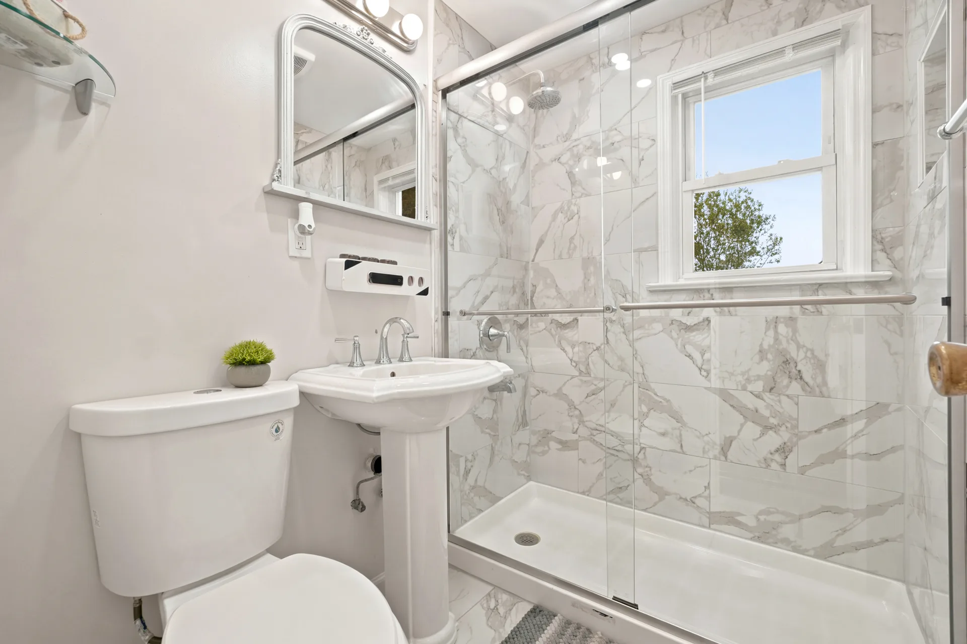 Renovated bathroom with white pedestal sink and glass-enclosed shower with marble-look tile