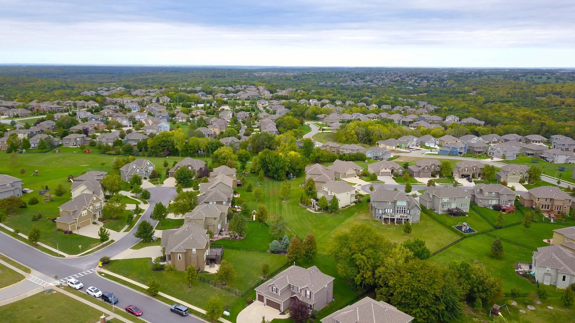 Aerial view of a sprawling suburban residential neighborhood with single-family homes, winding streets, green lawns, and mature trees under an overcast sky
