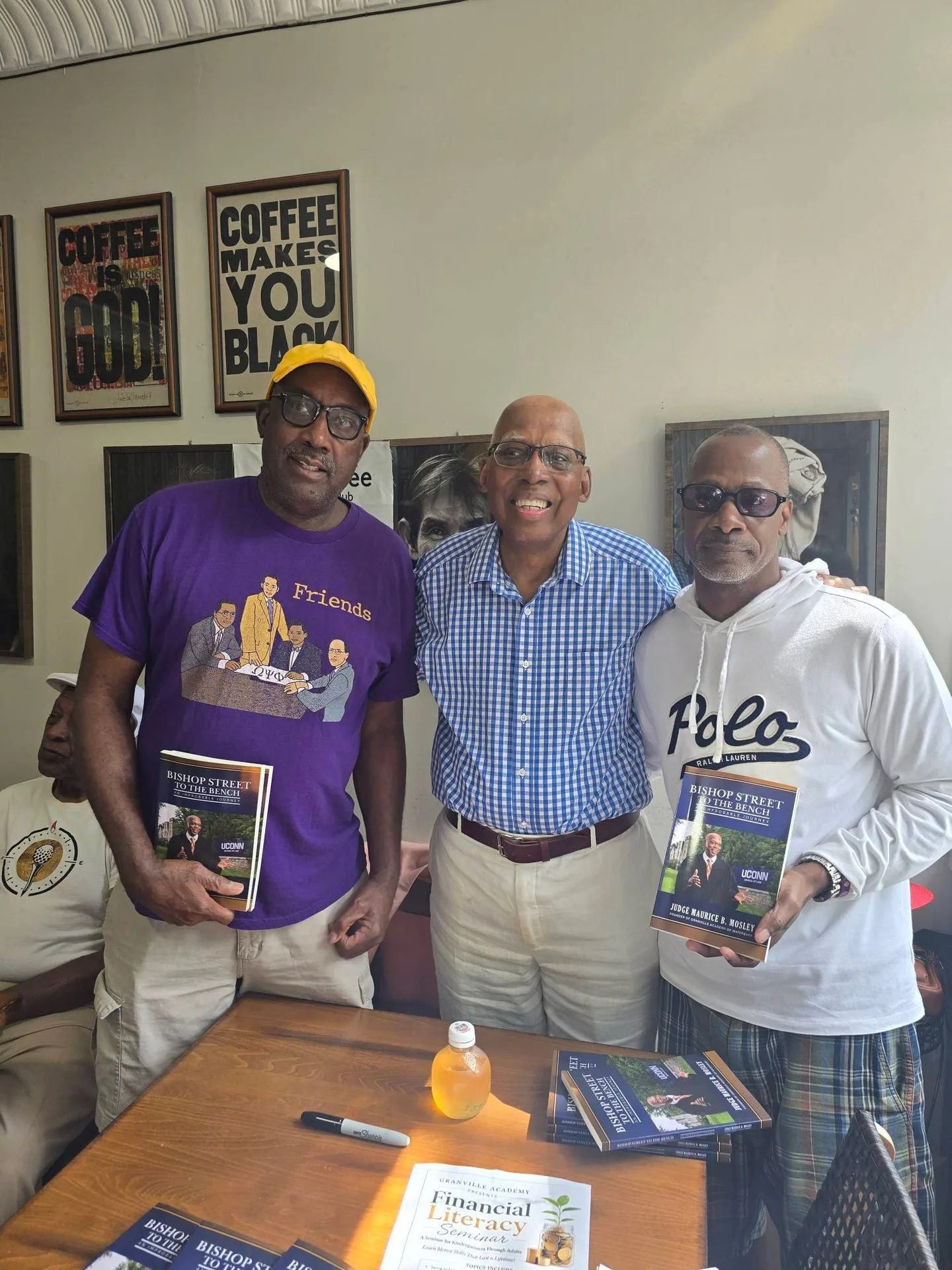 Three men standing behind a table inside the cafe holding copies of Bishop Street to the Pulpit, with copies of the book stacked on the table