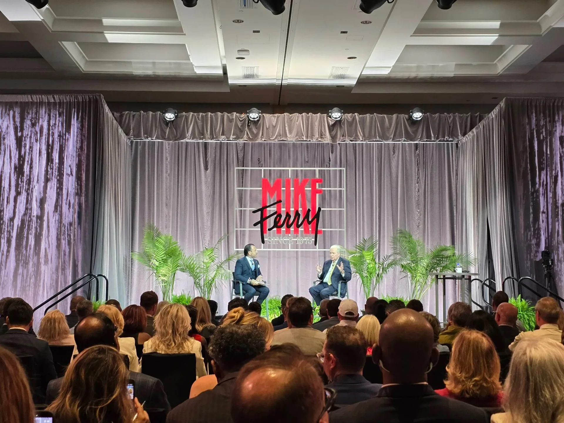 A fellow attendee, DK, seated on stage in conversation with Mike Ferry, who has white hair and wears a suit, the two facing each other in chairs with the MIKE FERRY backdrop behind them and the audience seated in front at the Mike Ferry 2026 Superstar Retreat at The Diplomat Beach Resort in Hollywood, Florida