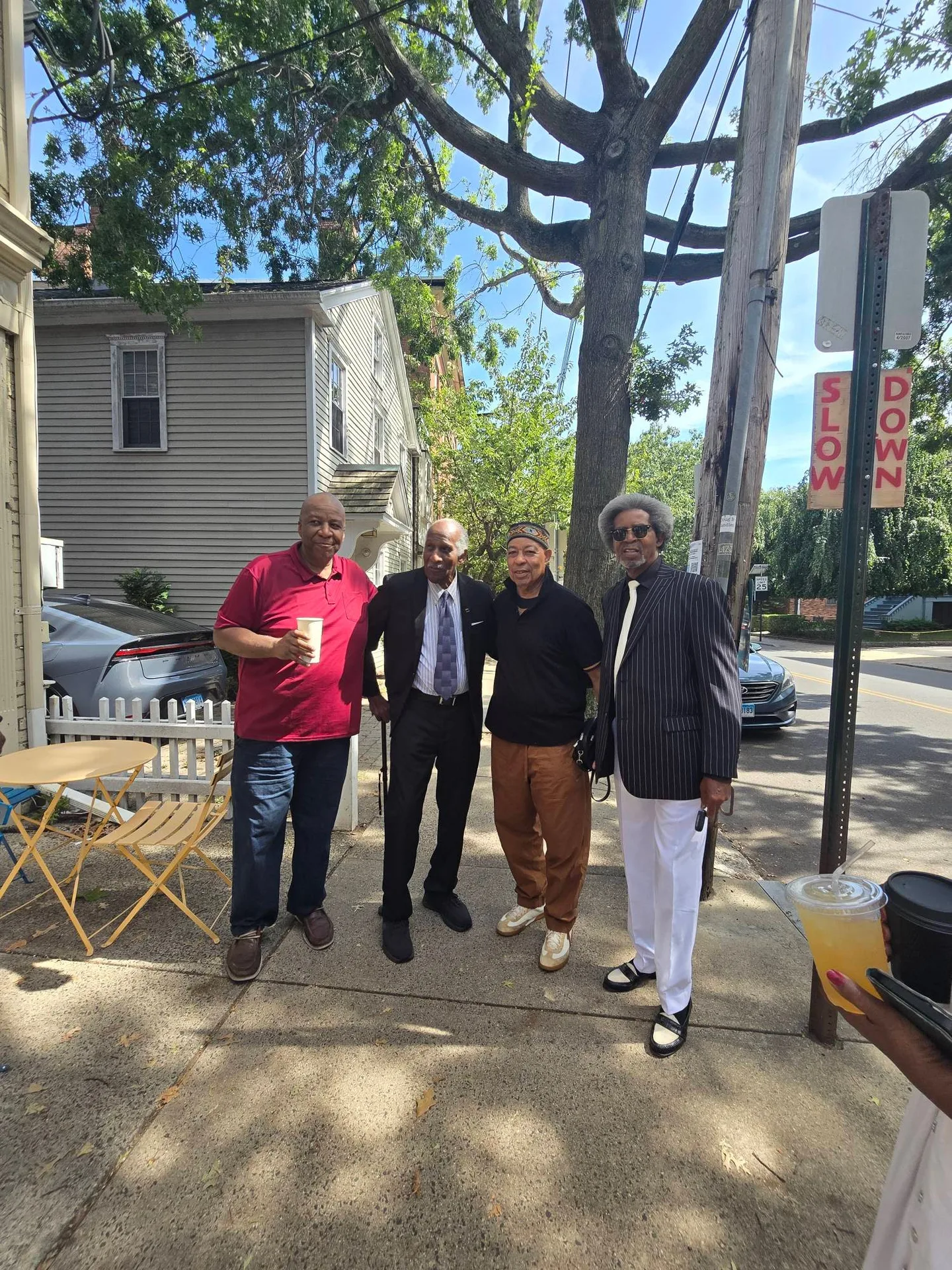 Four men gathered outdoors on the sidewalk near the cafe, with a SLOW DOWN sign visible