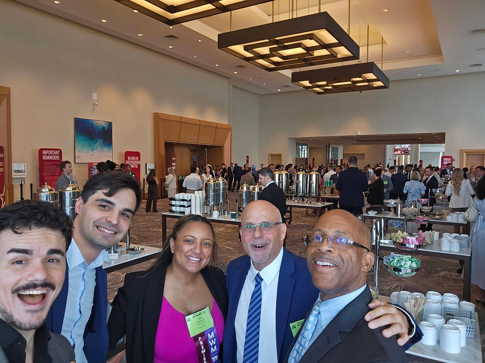 A group selfie of five attendees at the Mike Ferry 2026 Superstar Retreat at The Diplomat Beach Resort in Hollywood, Florida, smiling together with conference name badges during a break, with buffet tables and coffee urns in the background