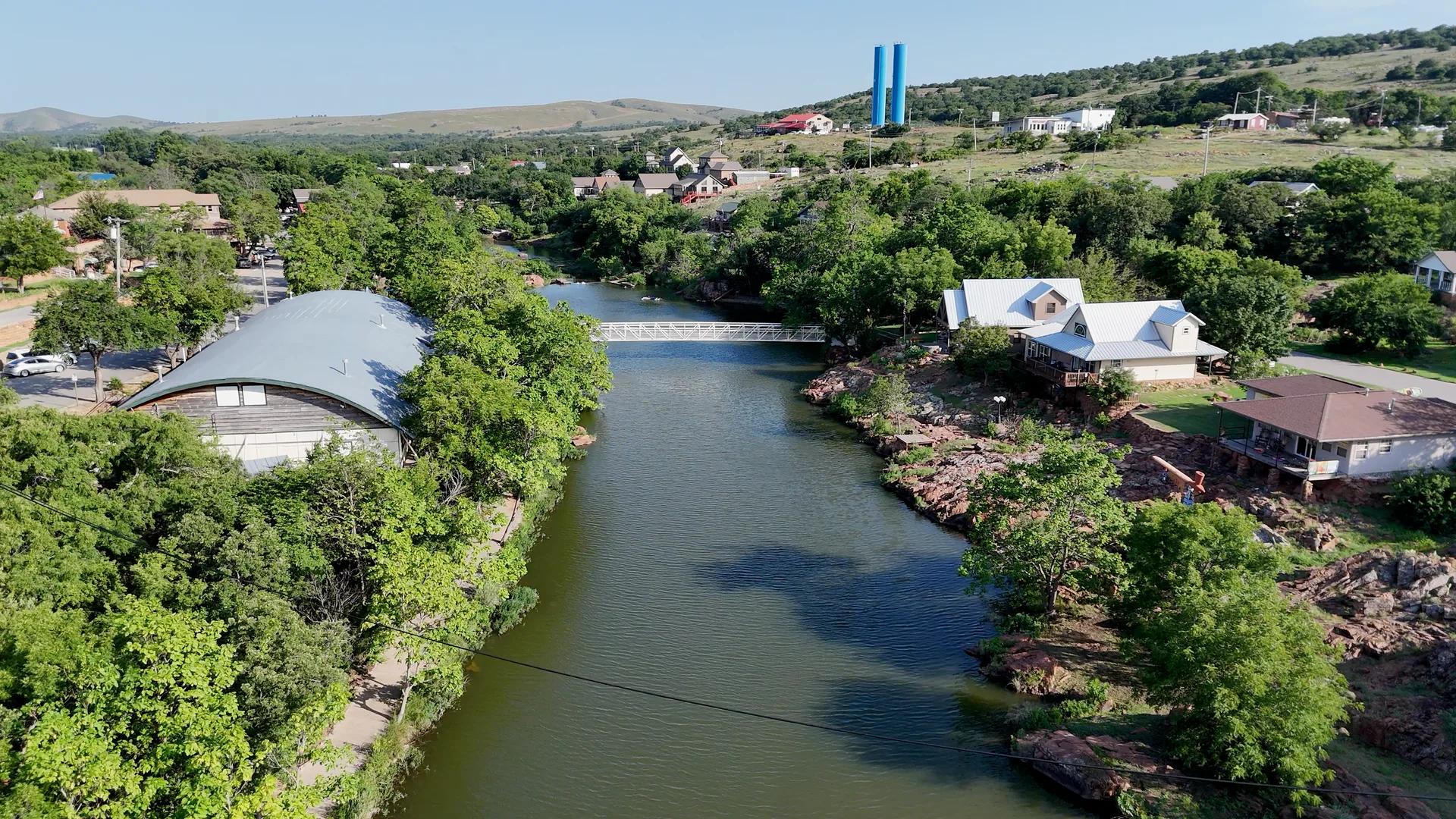 Aerial view of Medicine Park showing Bath Lake with its pedestrian footbridge, cottage-style architecture, and the two blue water towers