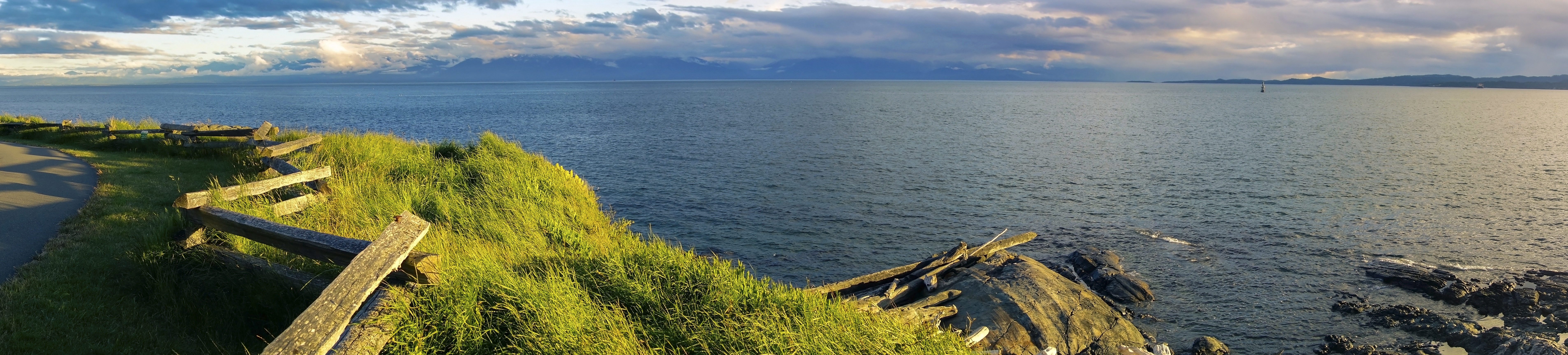 Panoramic view from Clover Point Park over the Strait of Juan de Fuca toward the Olympic Mountains, grassy cliffside, split-rail fence, dramatic sky