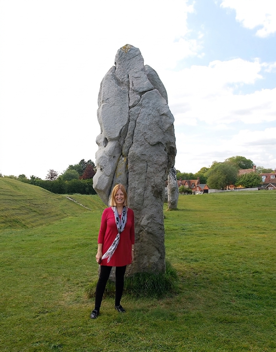Carol standing beside a tall stone at Avebury, wearing a red tunic and a patterned scarf.