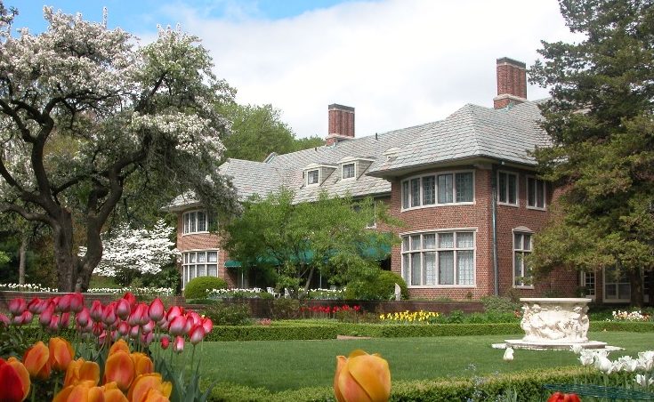 The brick and stone Tudor-Jacobethan manor house of Applewood Estate in Flint, Michigan, set among manicured lawns, a perennial garden, and an heirloom apple orchard in warm late-afternoon light