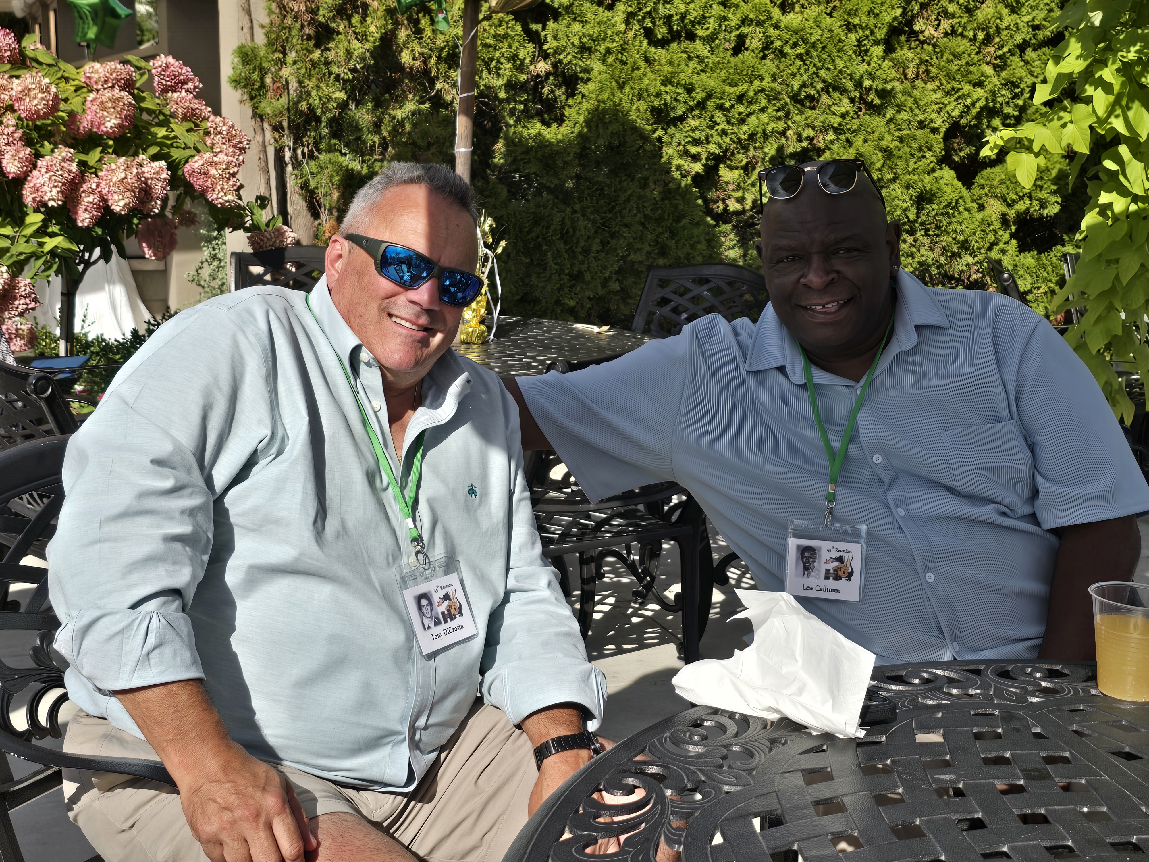 Two men in light blue button-down shirts and sunglasses, wearing green lanyards with reunion name tags, sitting together at a patio table at the Hamden High Class of 1981 45th reunion.