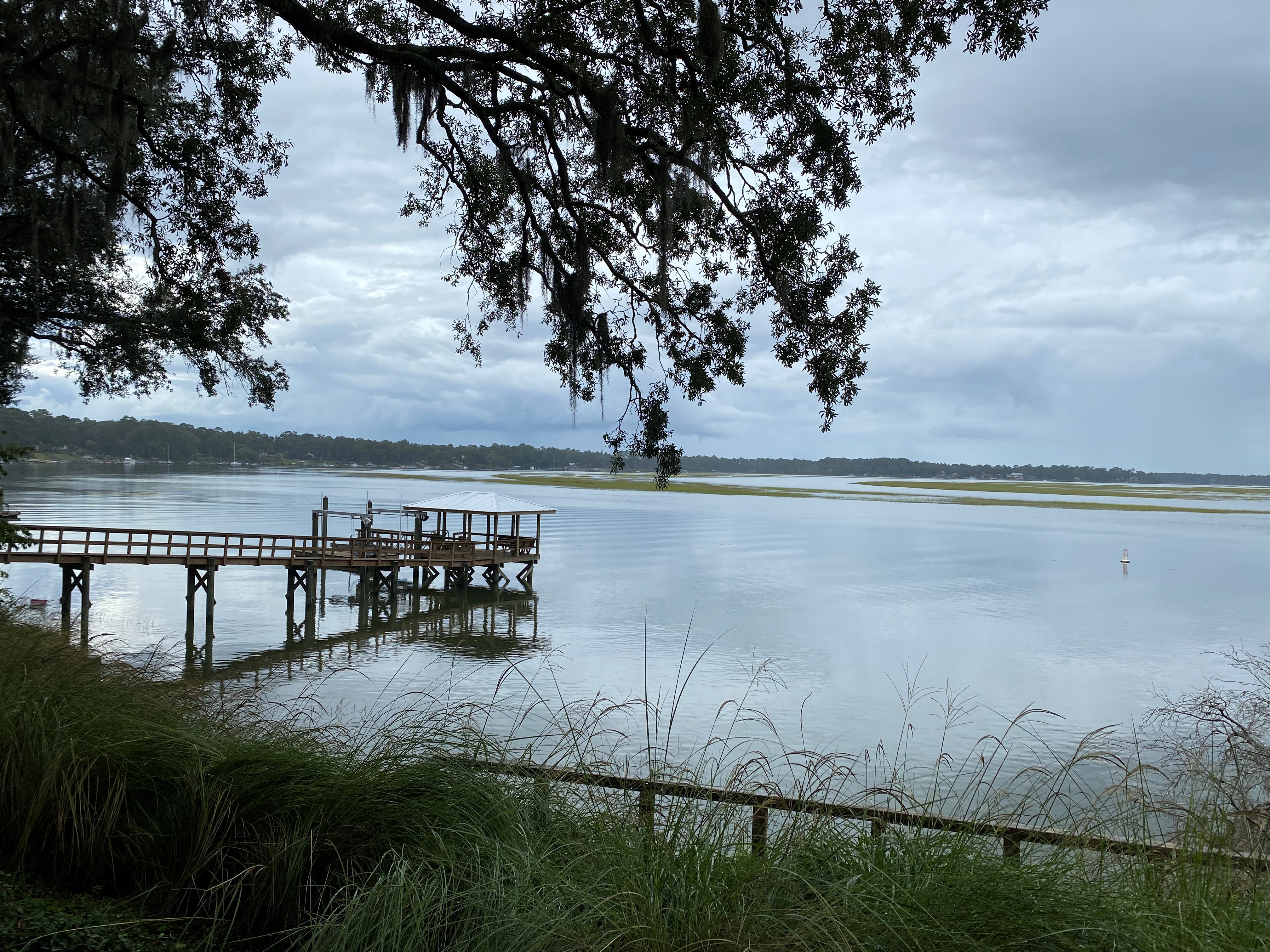 Overcast view of the May River, green marsh grasses in the foreground and a wooden pier with a covered platform extending into calm water, a low marsh island on the horizon under muted greens and cool desaturated blues and greys